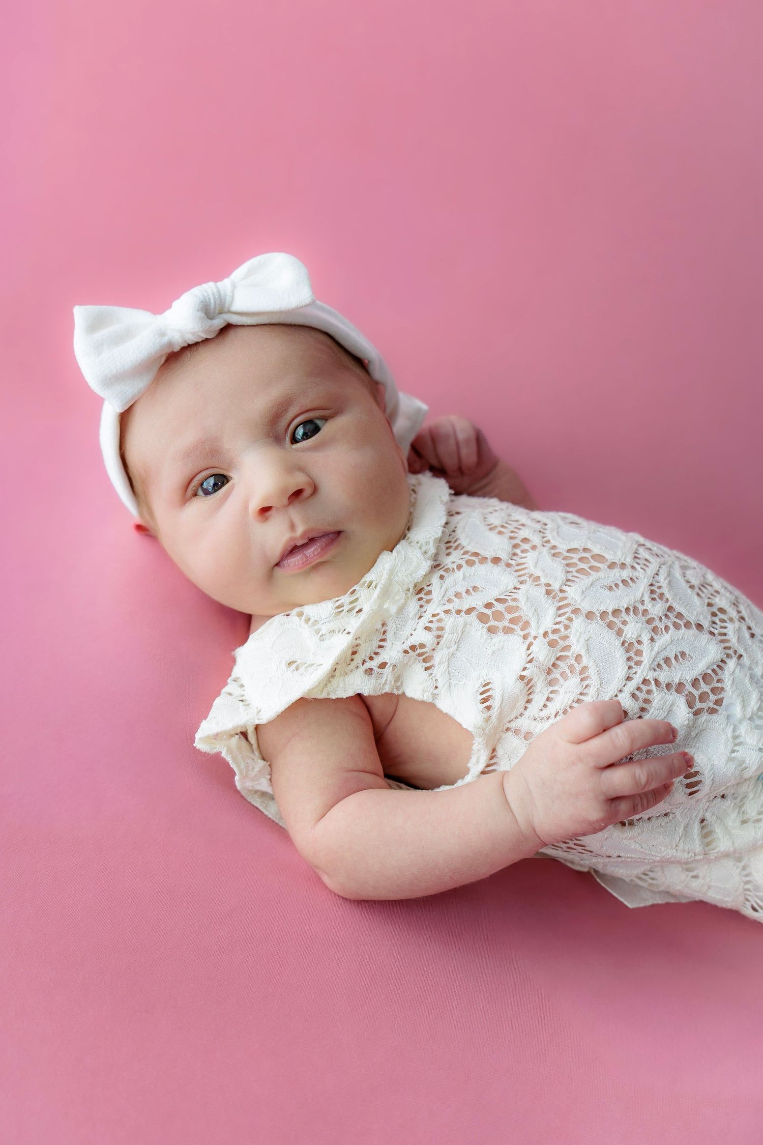 A newborn baby wearing white lace dress and cap lies on pink background in a professional portrait series.