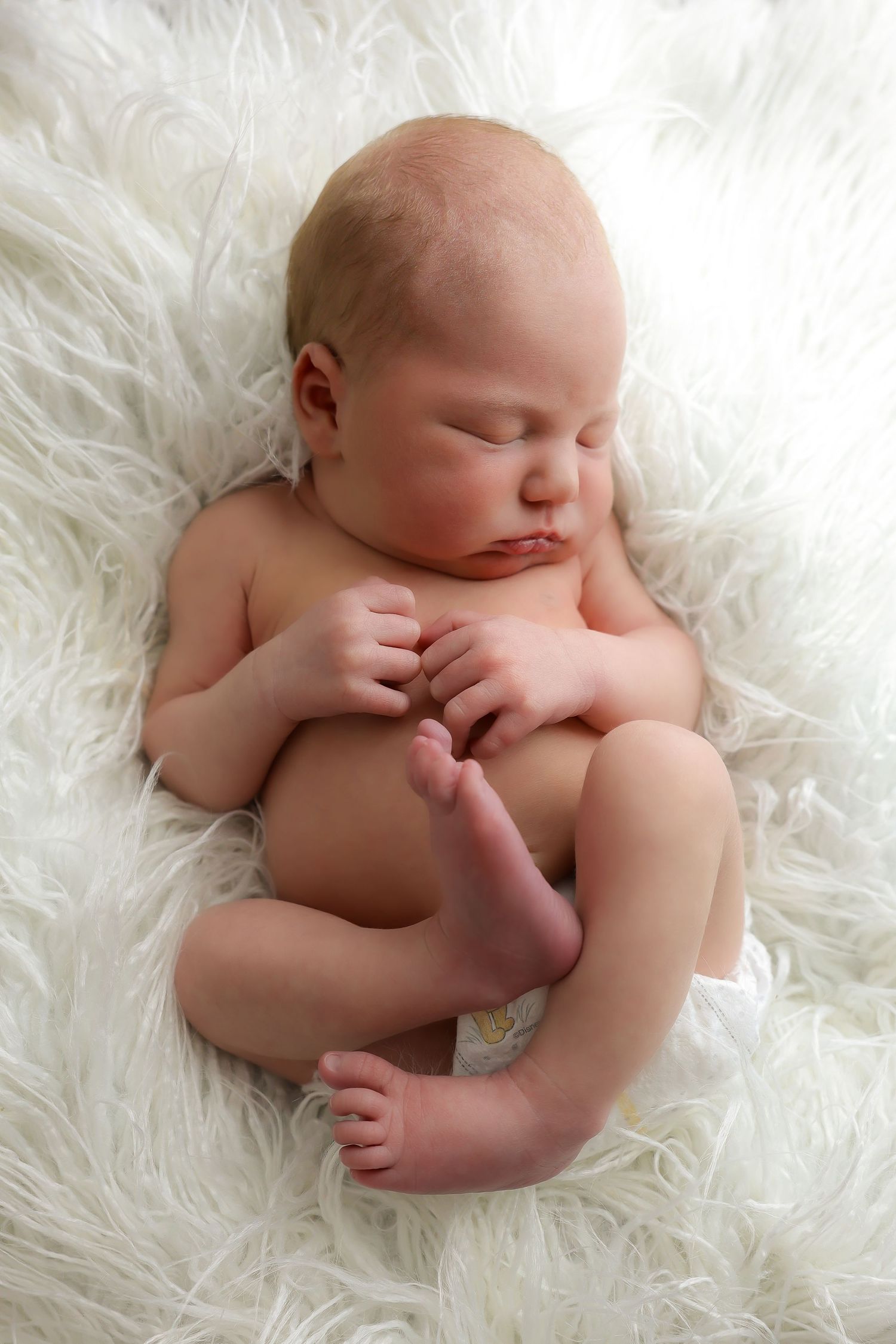 Peaceful sleeping newborn curled up on soft white furry blanket in natural light.