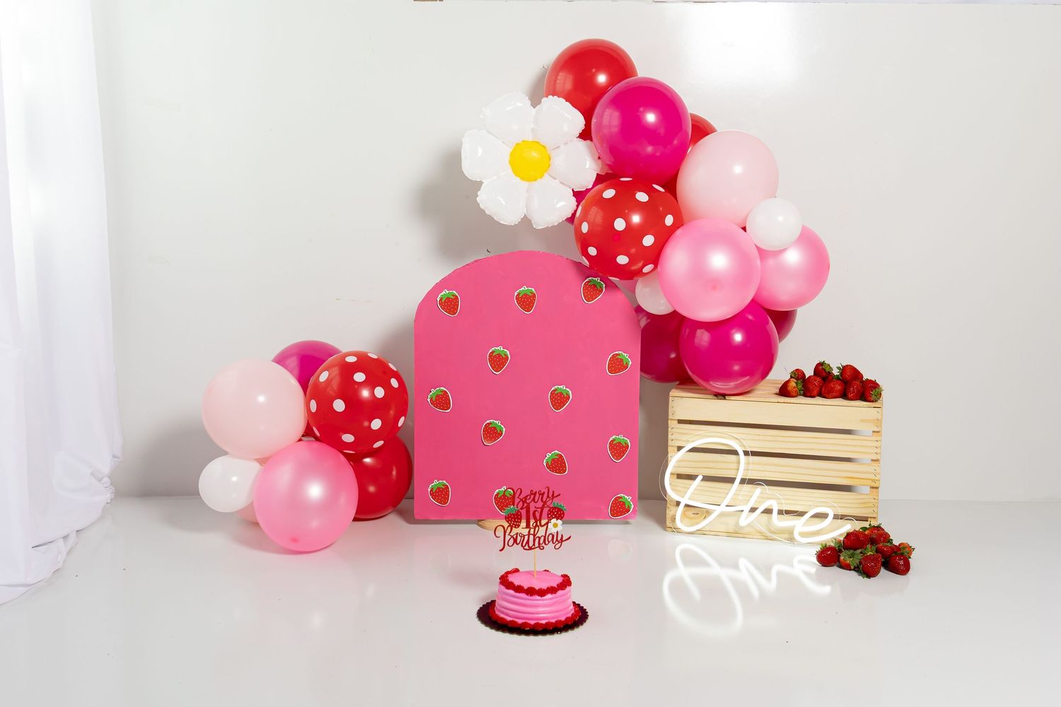 Whimsical pink and red party setup with balloons and wooden crate against white background.
