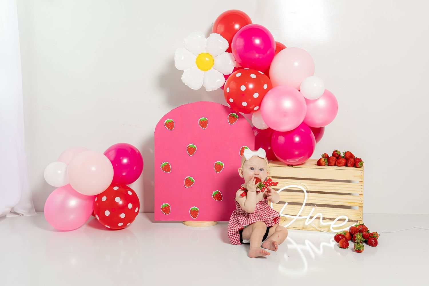 Party setup with pink balloons, wooden crate, and floral decorations against white wall.