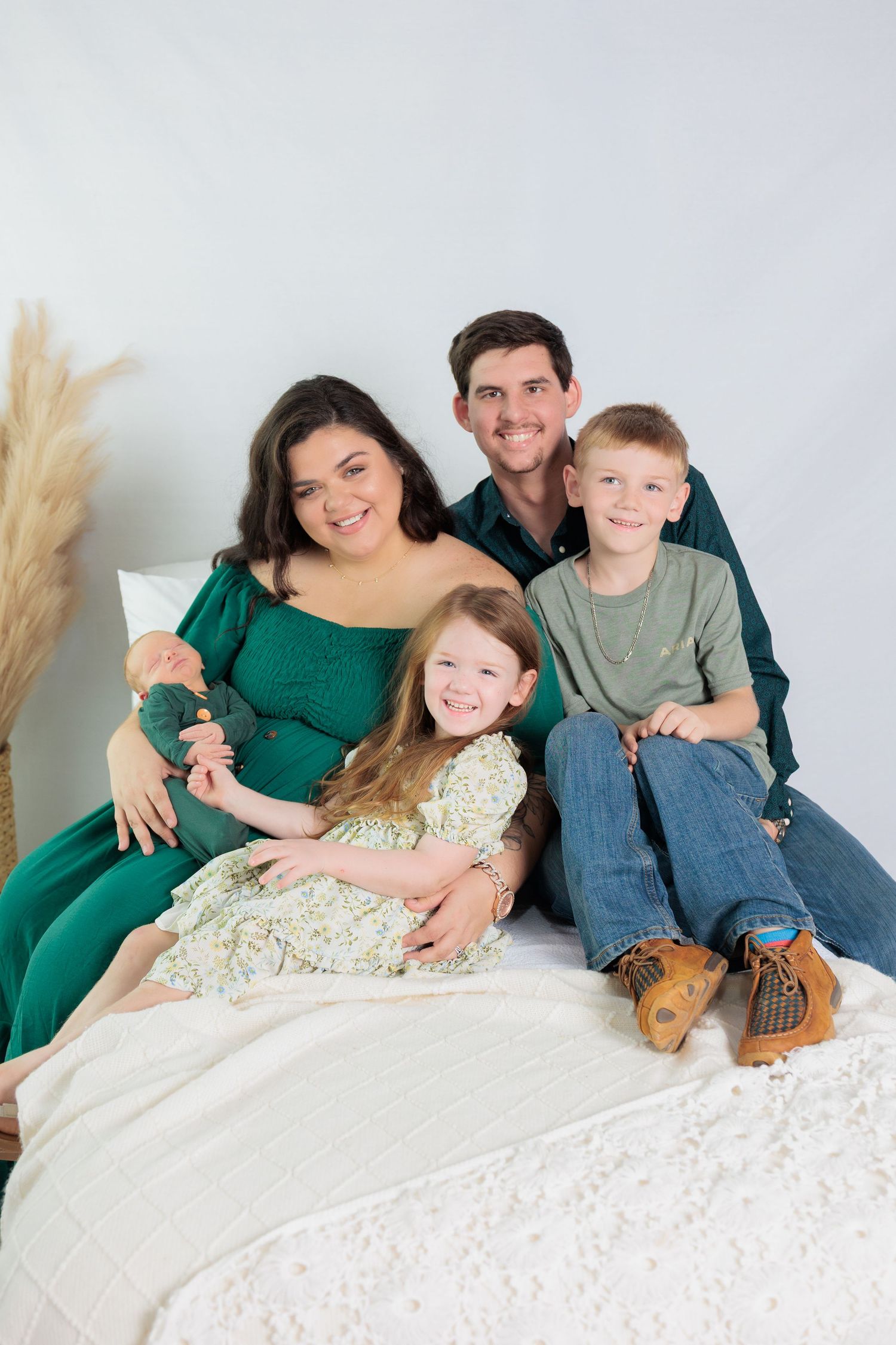 A family in coordinating green and blue outfits poses together on a white bed with decorative pampas grass.