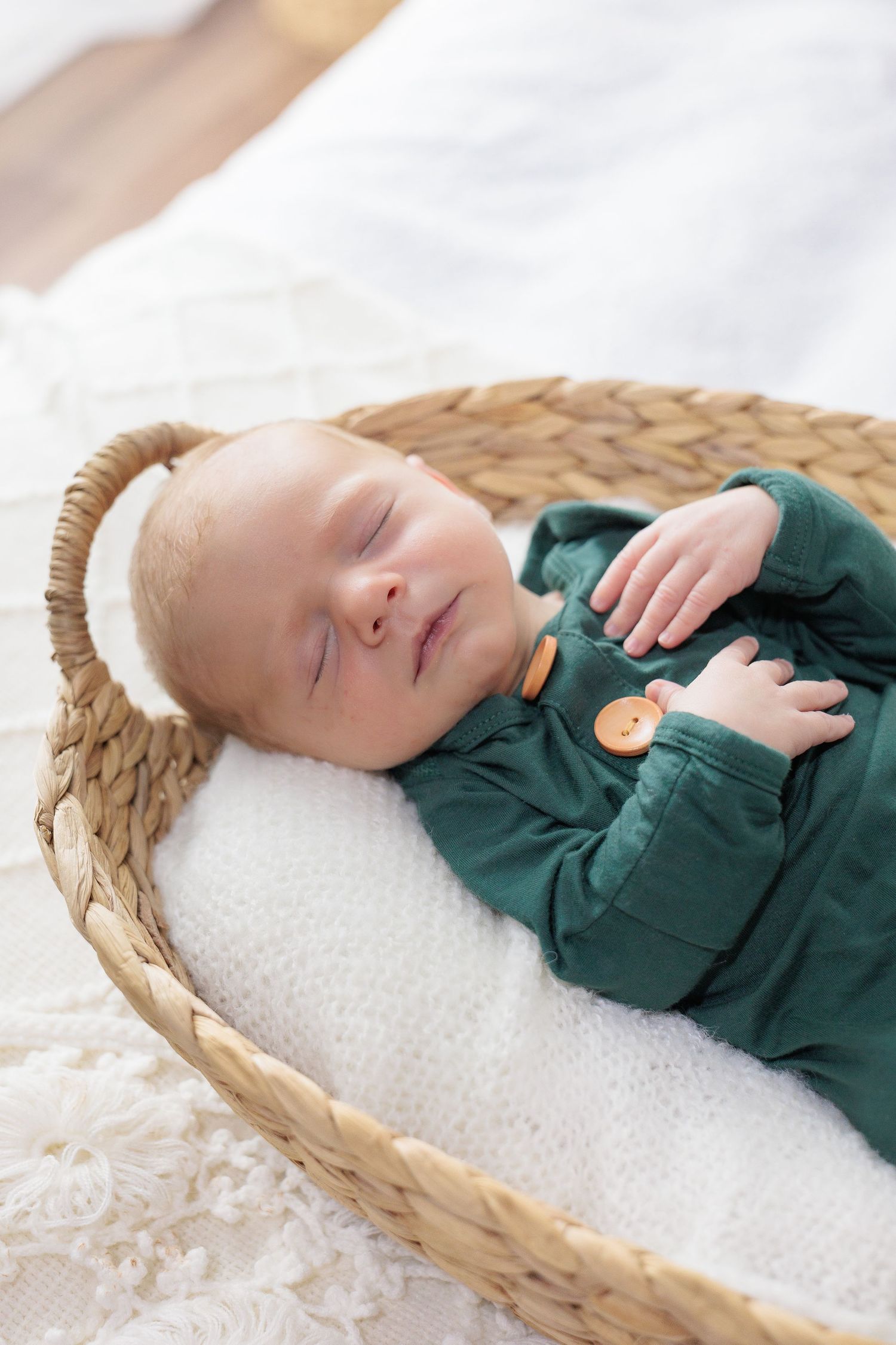 Sleeping baby wearing green outfit rests peacefully in woven basket on white blanket.
