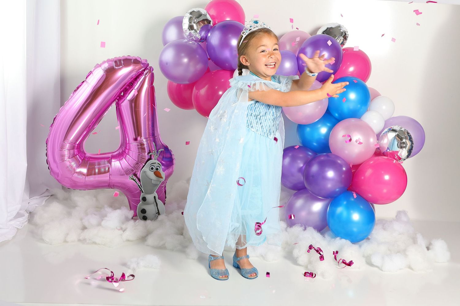 A child in a blue dress poses with colorful pink and purple balloons during a birthday photo session.