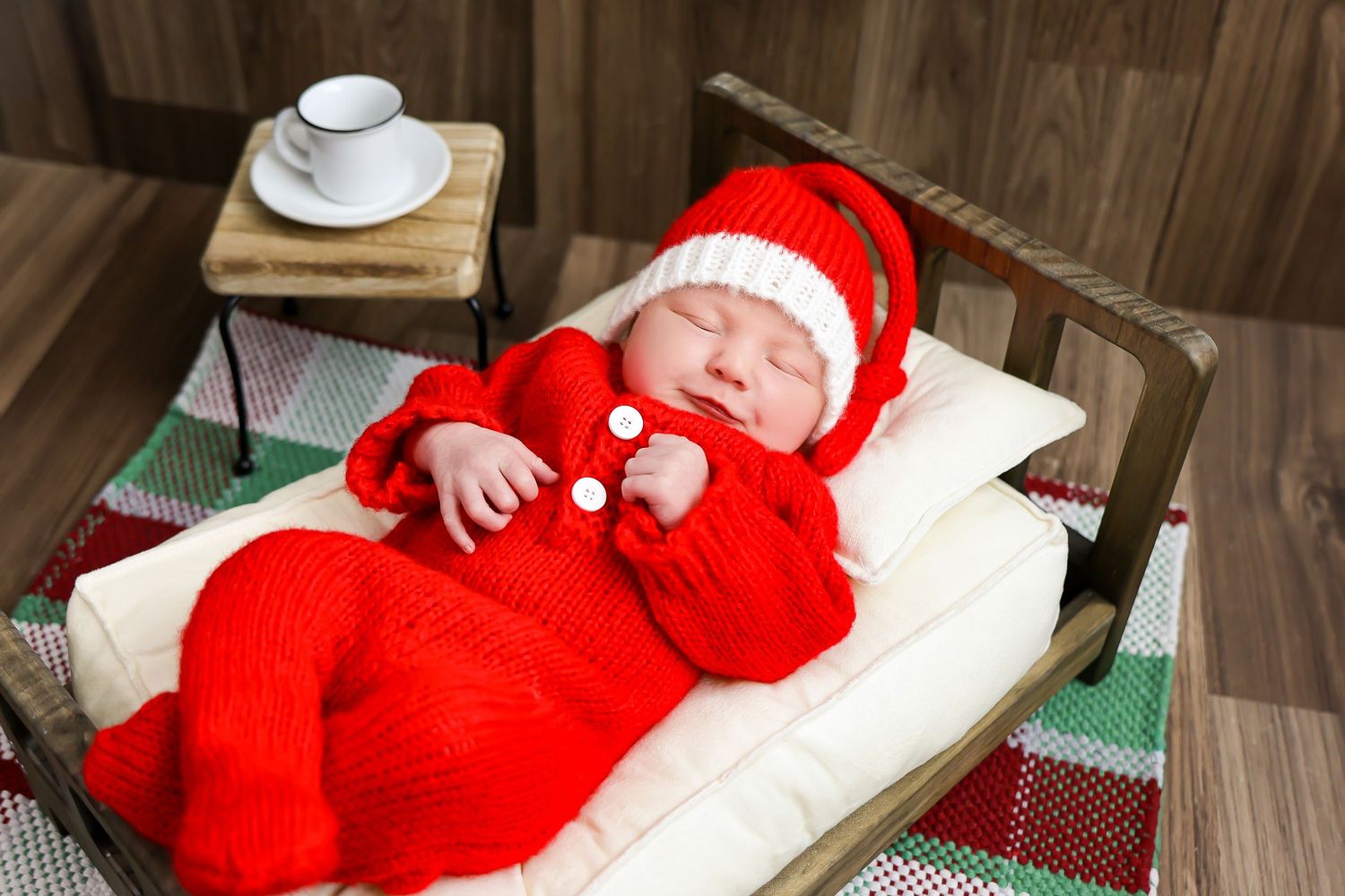 A sleeping baby wearing a bright red Santa outfit lies on a white cushion during a Christmas photo session.