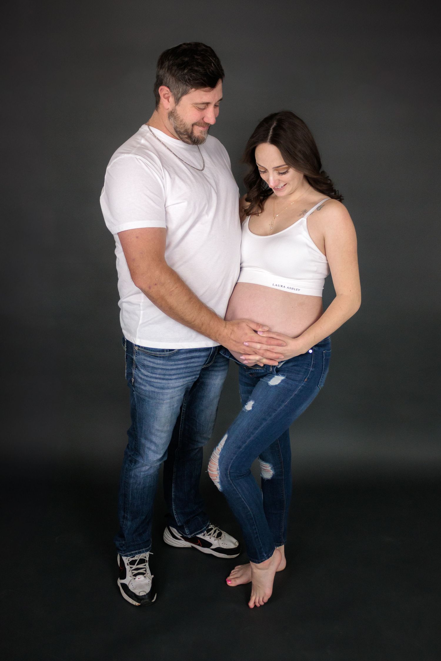 Casual maternity photos of couple in jeans and white tops on dark background.