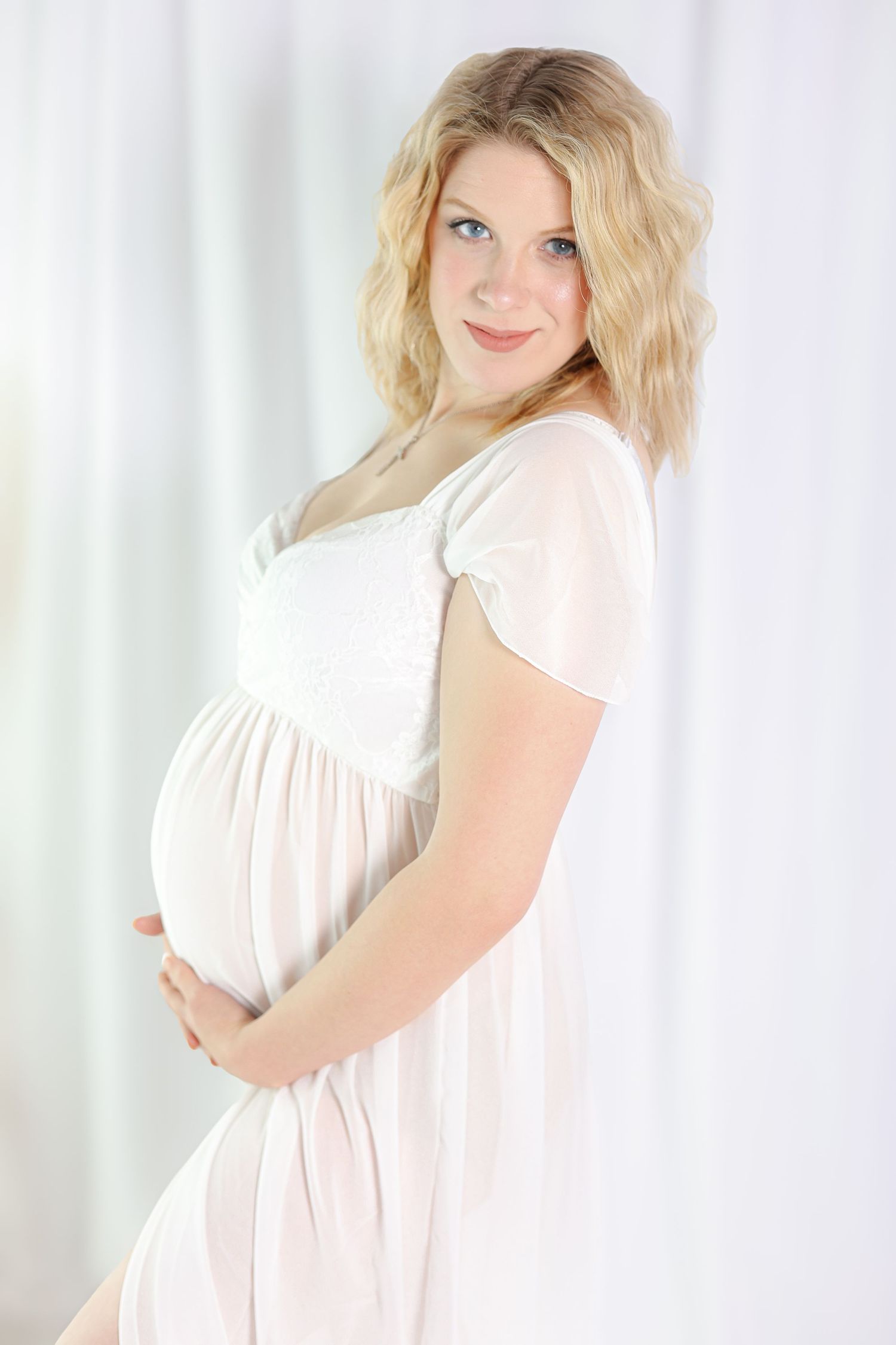 Maternity portrait in flowing white dress against light background.