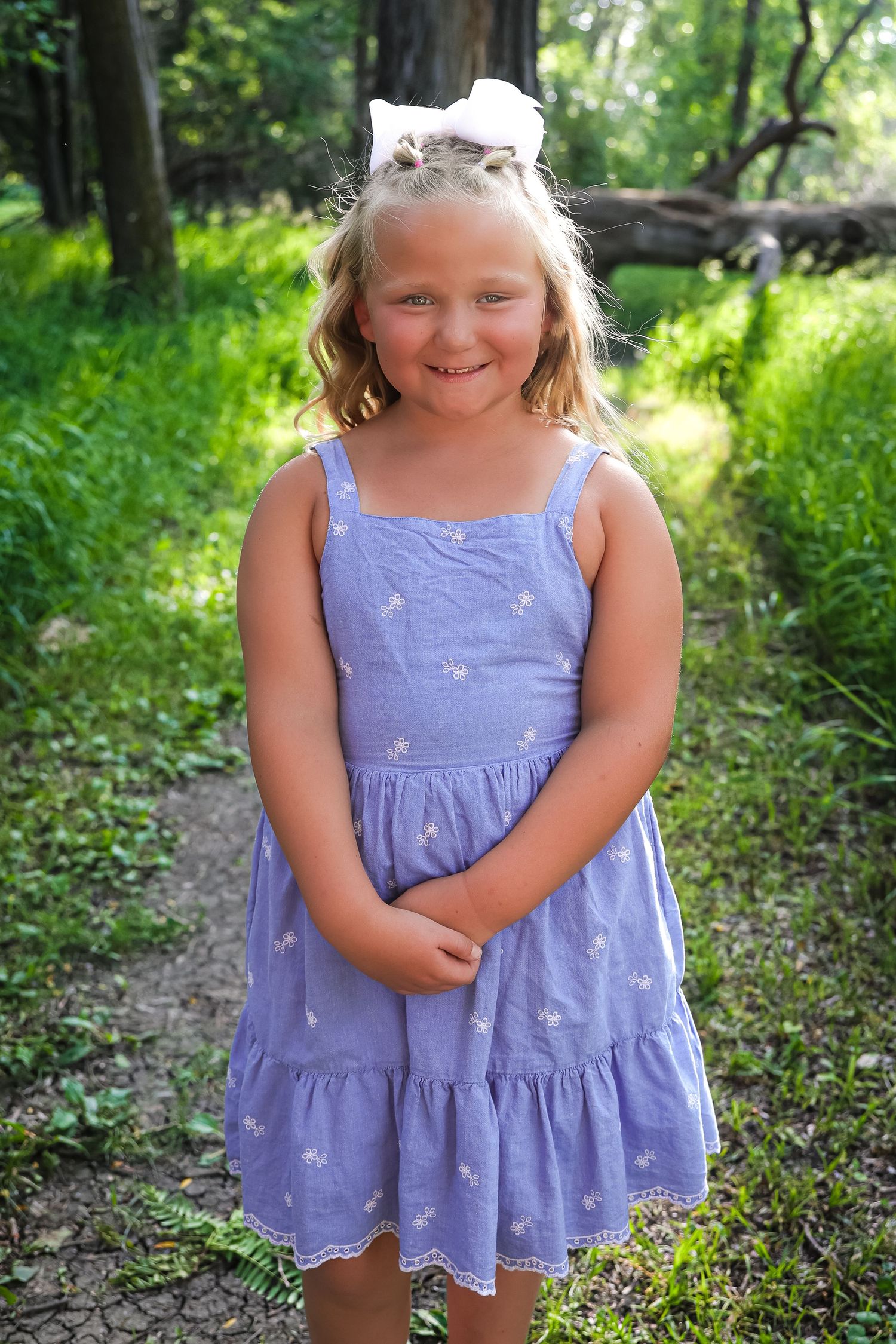 Child in purple summer dress smiles while posing in a wooded outdoor setting.