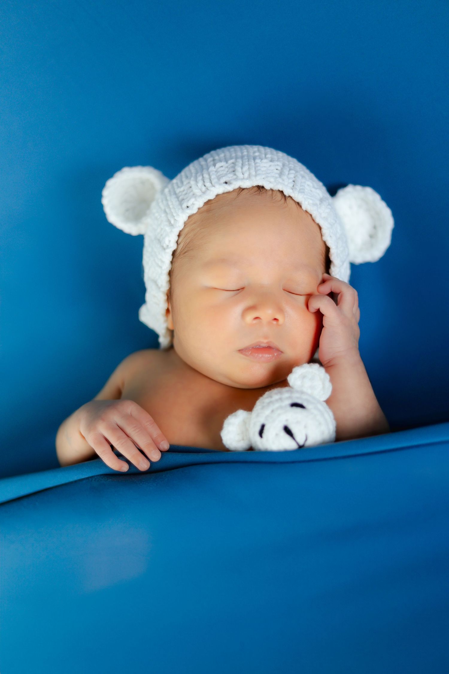Sleeping newborn in white knit hat with plush toy against blue backdrop.