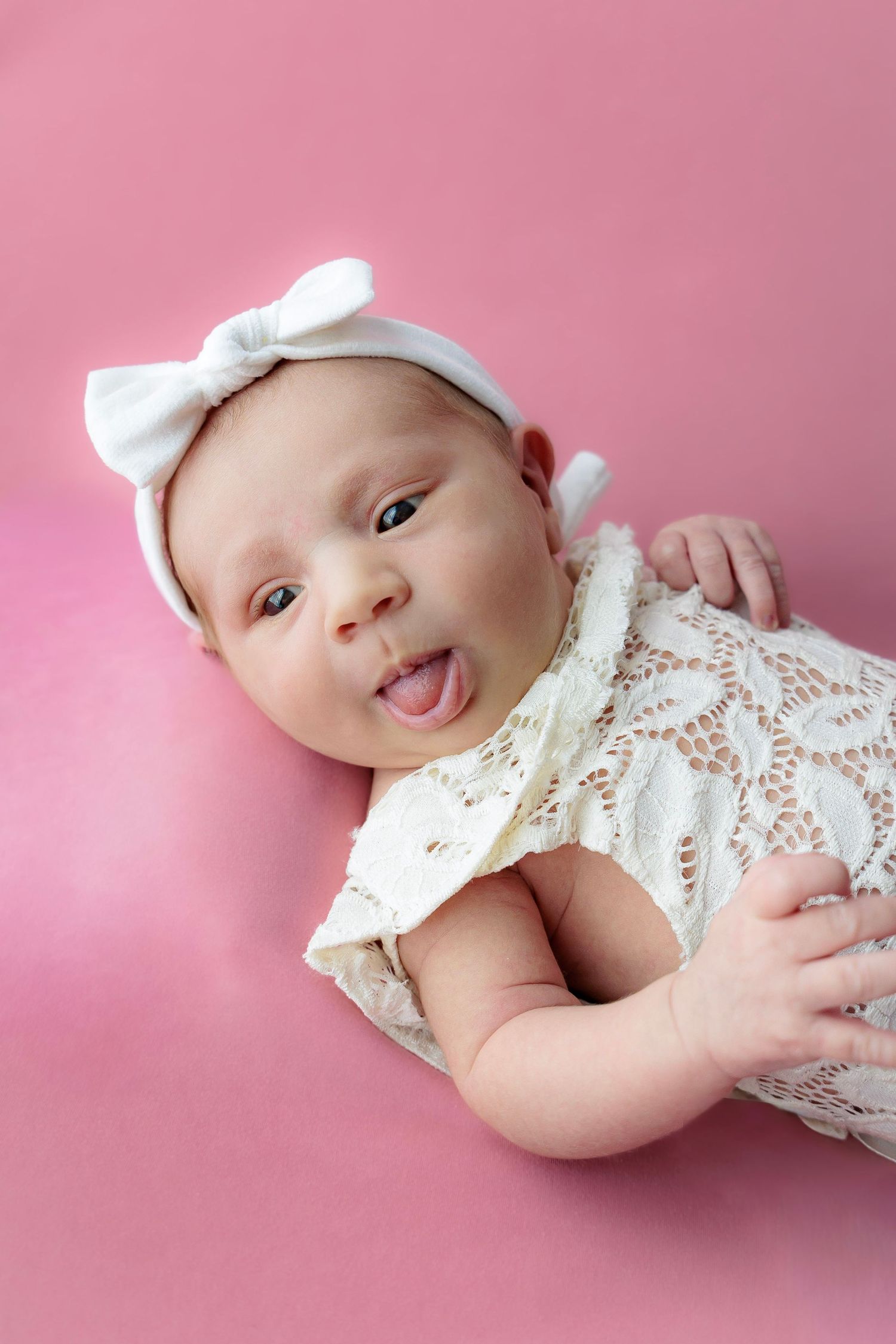 A baby wearing a white lace outfit and bow lies on a pink background while making sweet expressions.