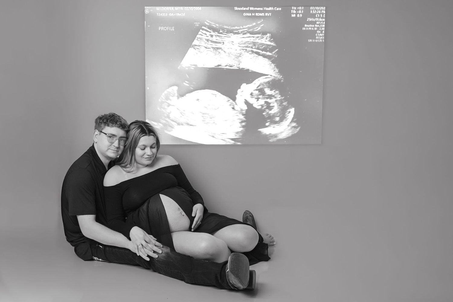 Black and white photo of an expectant couple sitting together with an ultrasound image projected on the wall.