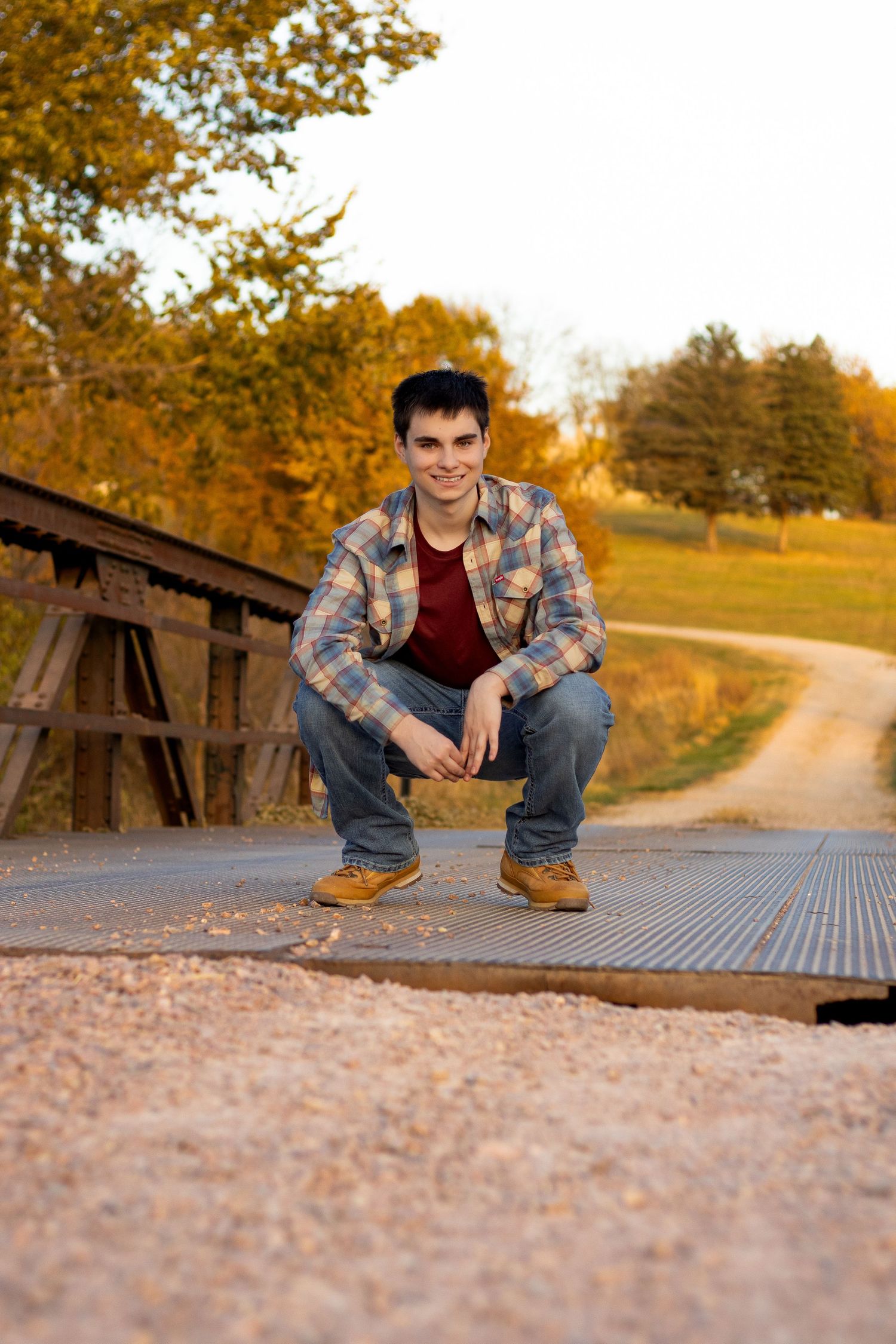 A person in plaid shirt and jeans crouches on a wooden bridge during autumn.