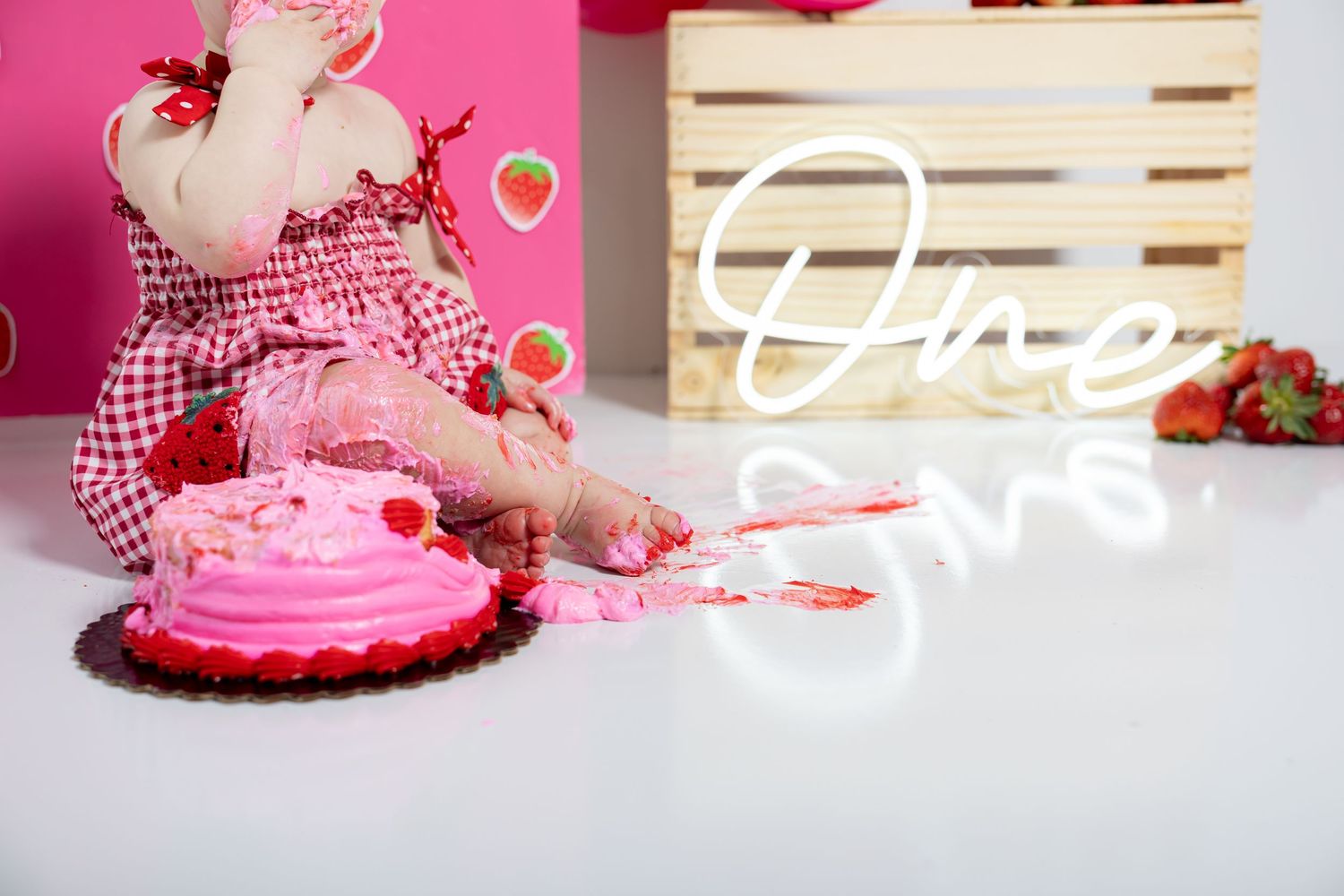 Pink cake smash session with gingham dress and strawberry decorations on white floor.