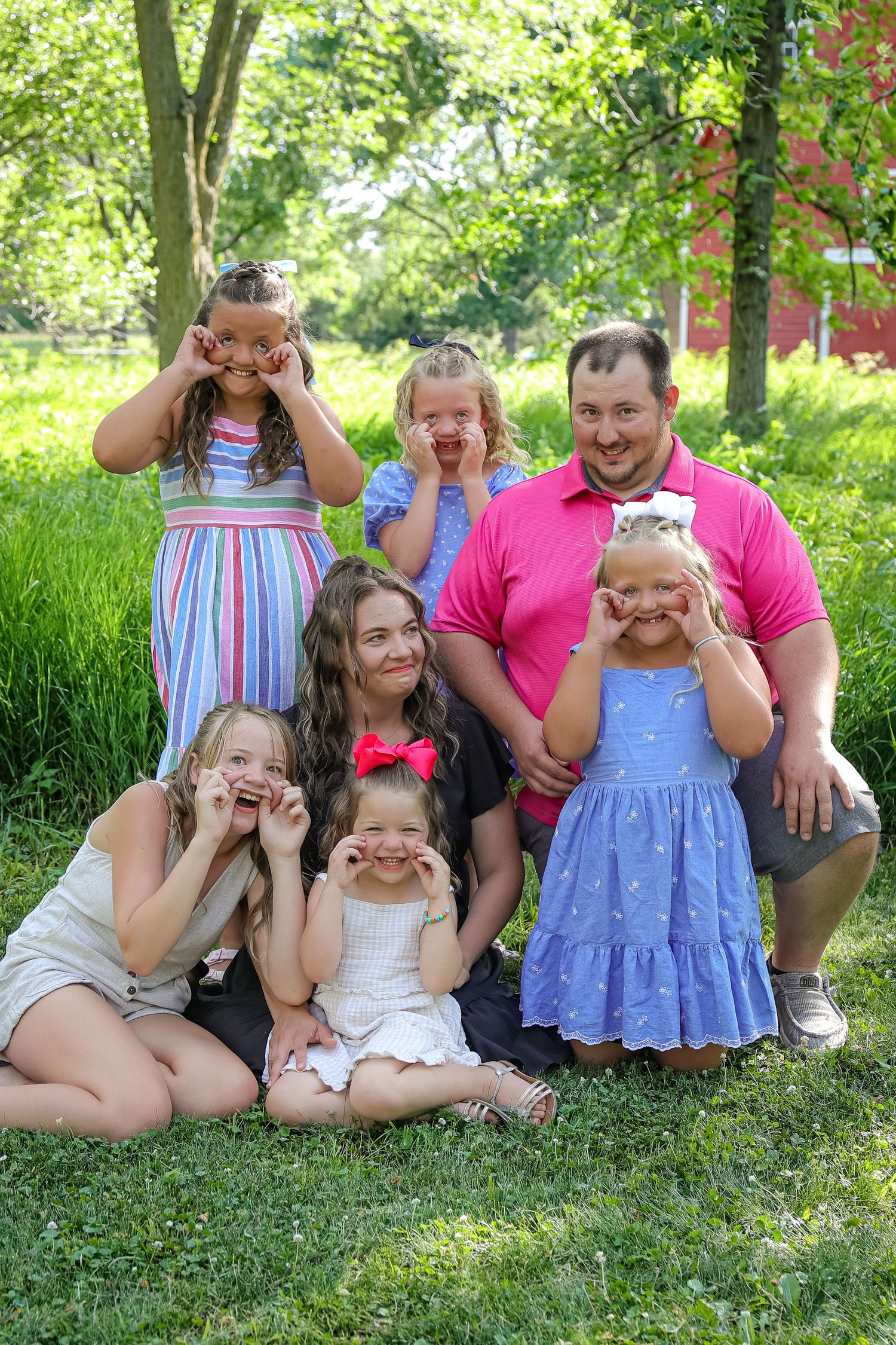 Family making playful expressions while sitting together on grass in a wooded setting.