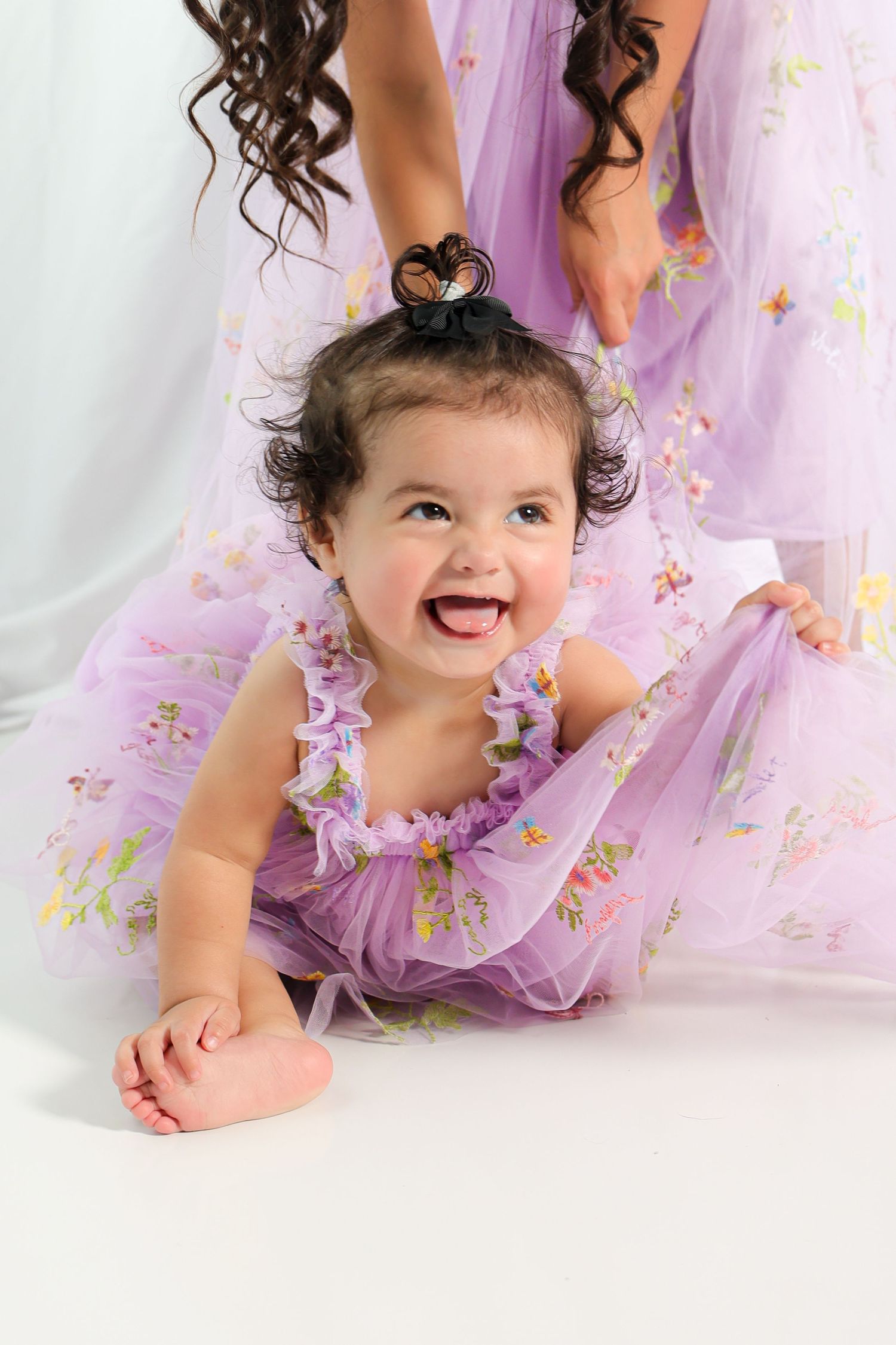 Baby in purple tulle dress smiles while lying on white floor with sparkly backdrop.