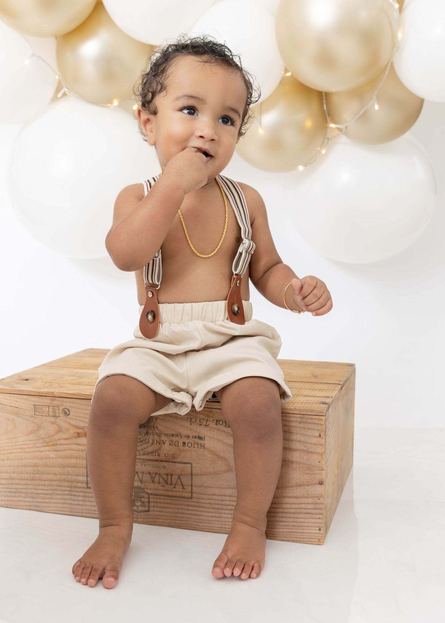 Baby in cream-colored romper sits on wooden crate against backdrop of gold and white balloons.