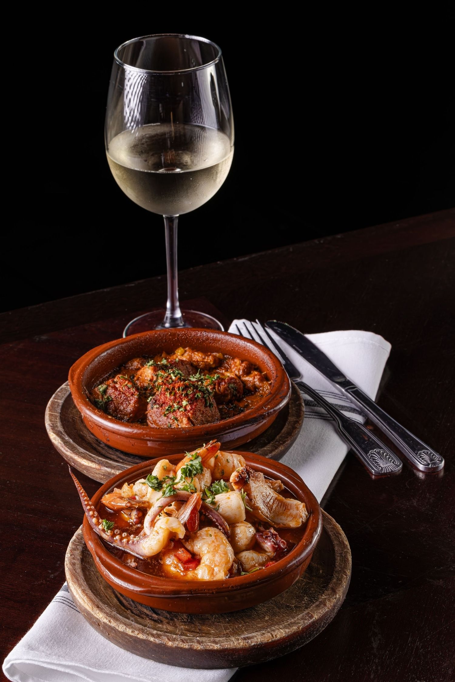 Spanish tapas dishes with meatballs in tomato sauce and seafood served with a glass of white wine on dark wooden table.