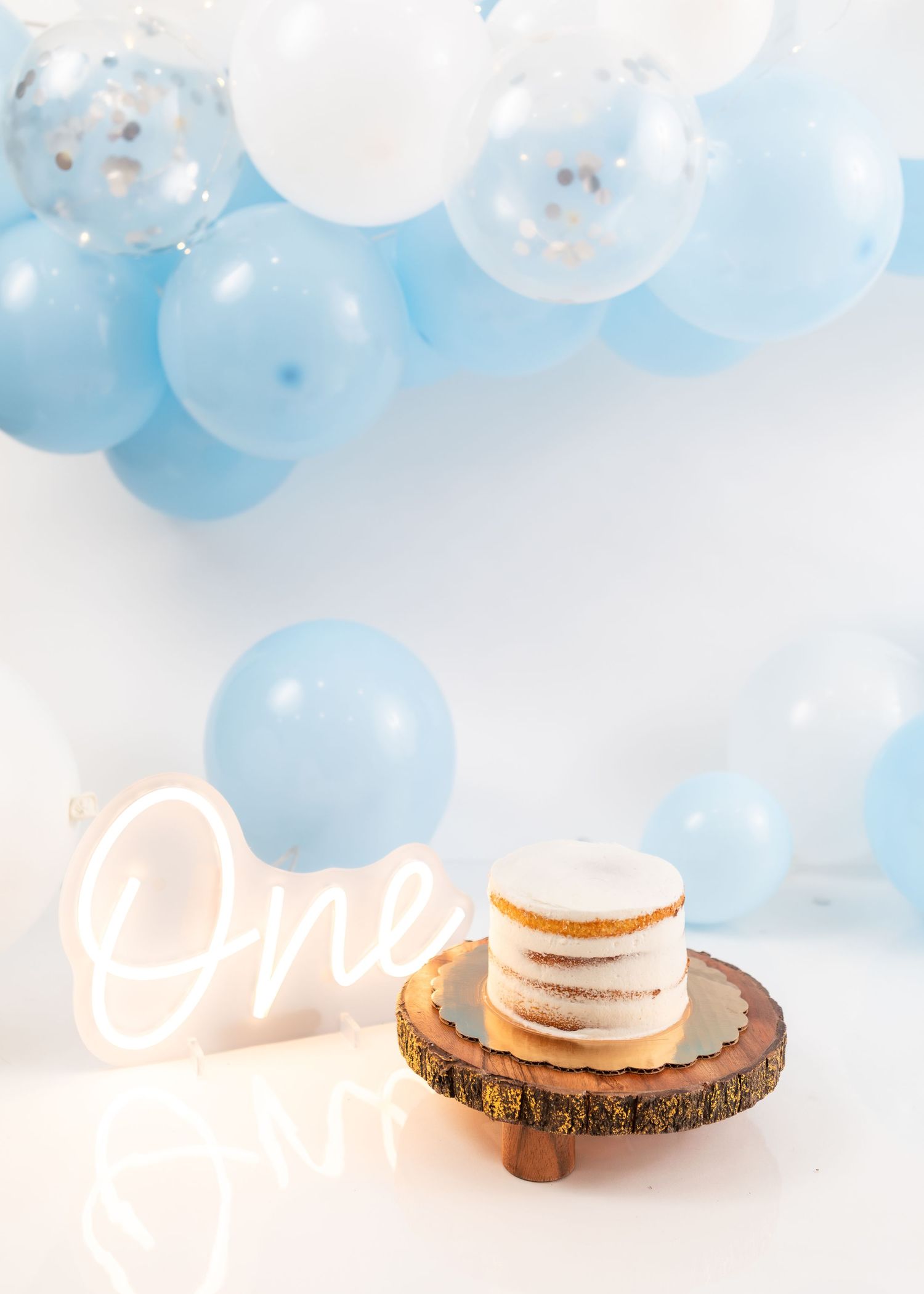 Minimalist cake smash setup with blue balloons and illuminated 'One' sign.