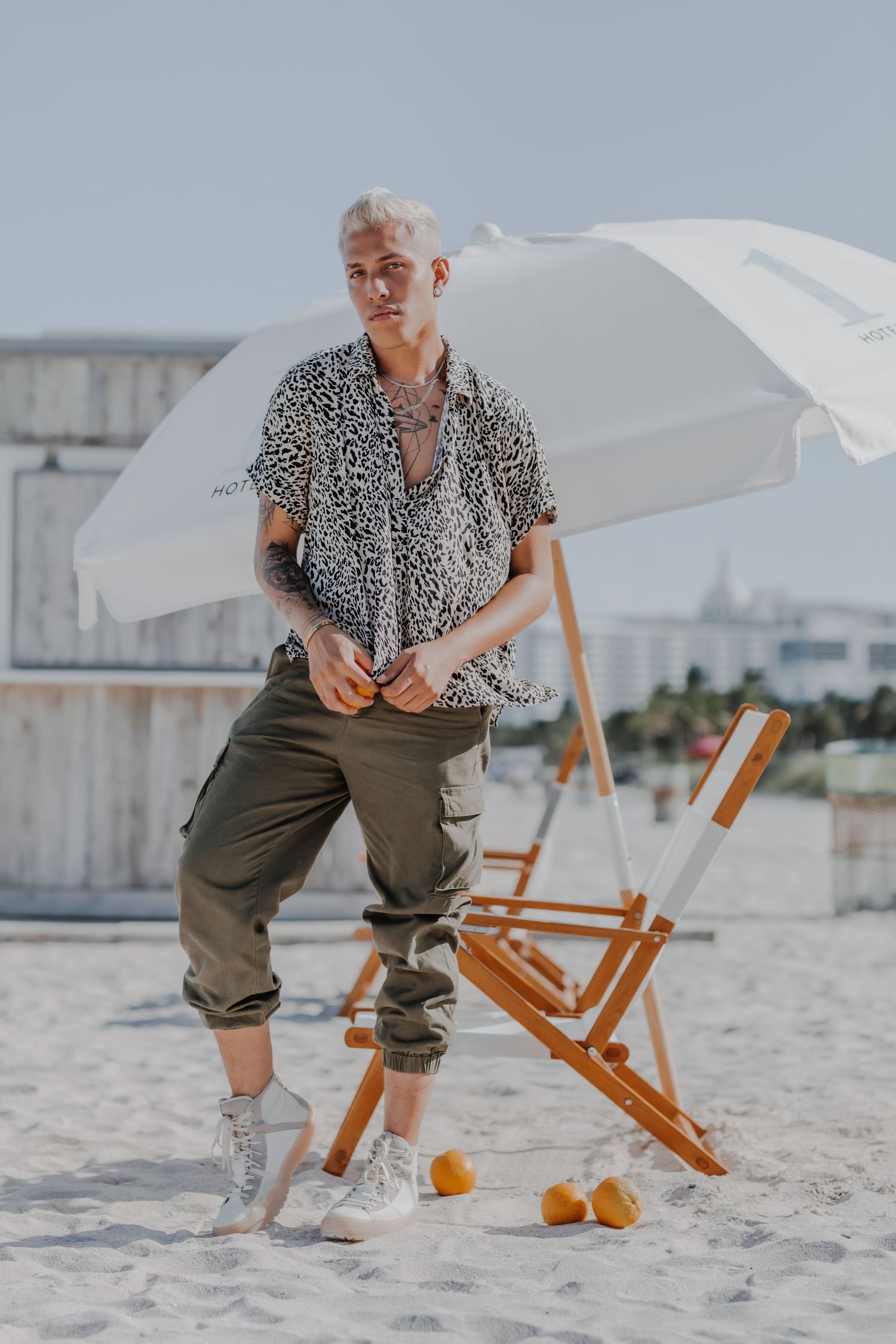 Stylish person in leopard print shirt and cargo pants posing on sandy beach with beach umbrella.