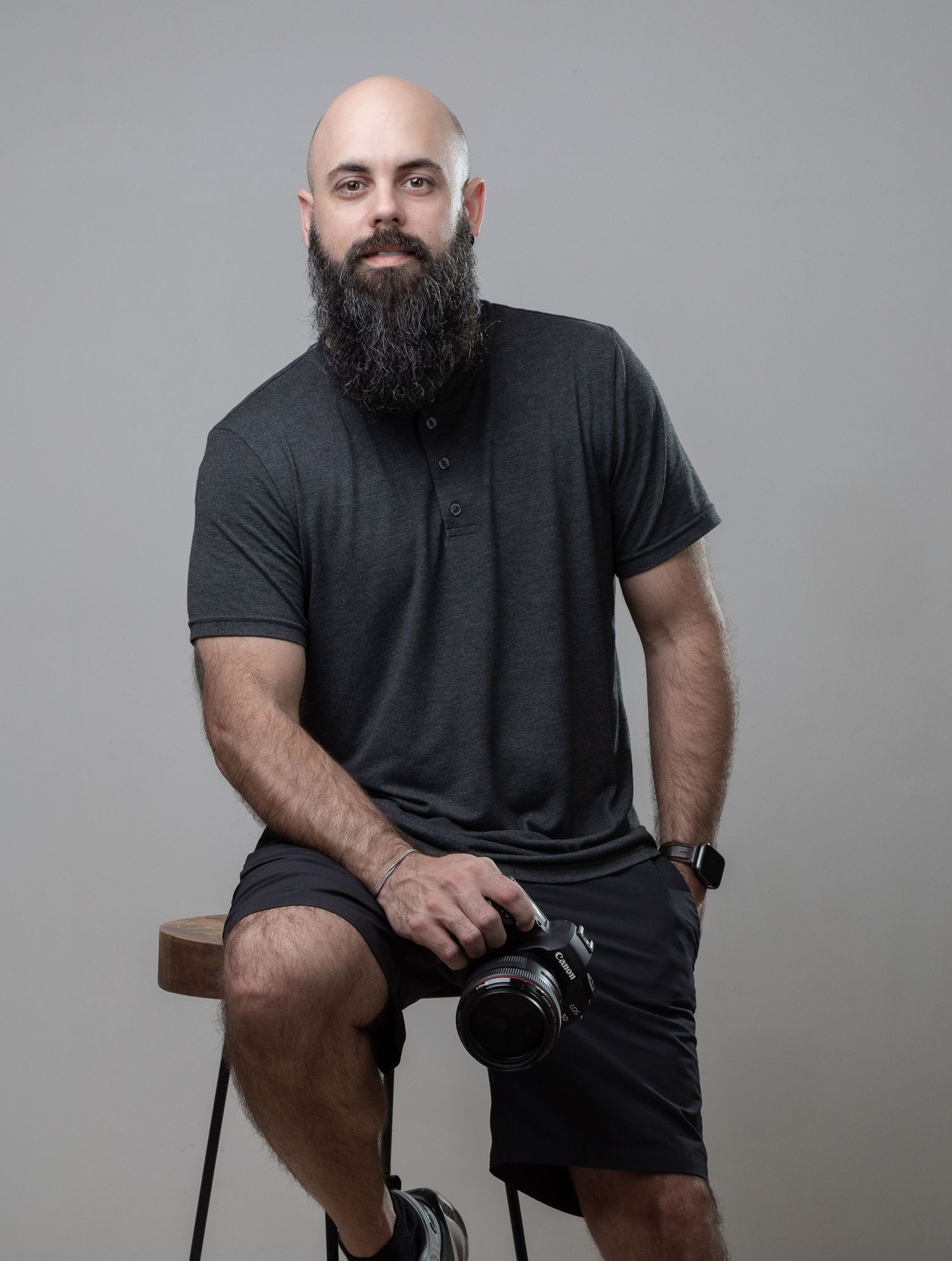 Professional photographer sitting on stool wearing dark t-shirt and shorts while holding camera in studio setting.