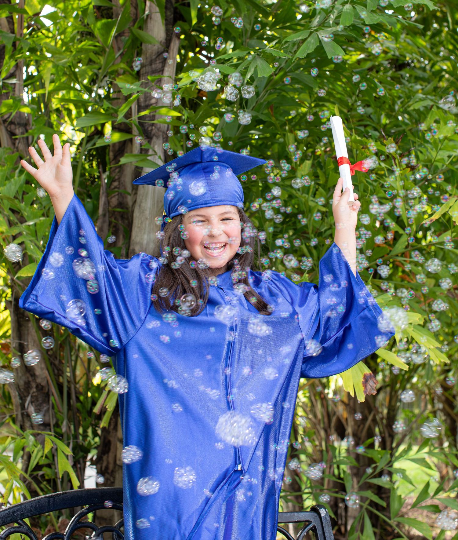 Graduate in bright blue cap and gown celebrates with diploma and raised arms in outdoor garden setting.