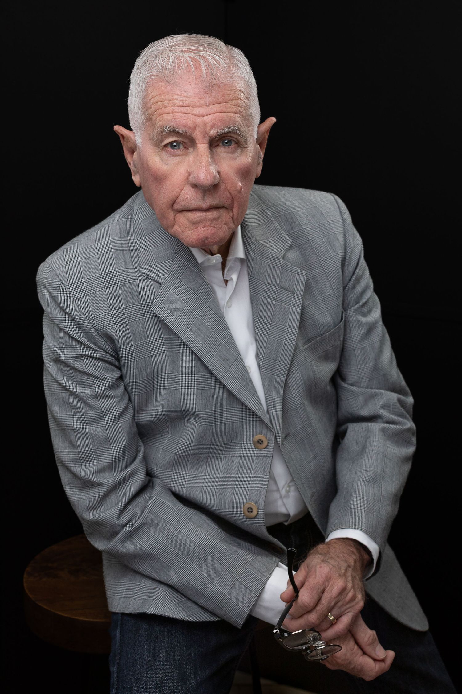Senior businessman in grey blazer seated for professional portrait against dark background.