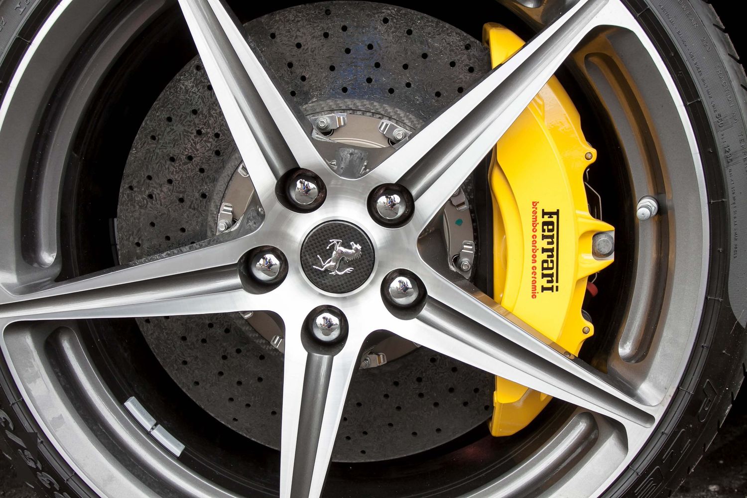 Close-up view of a Ferrari wheel with yellow brake caliper and drilled brake rotor behind a five-spoke silver wheel.