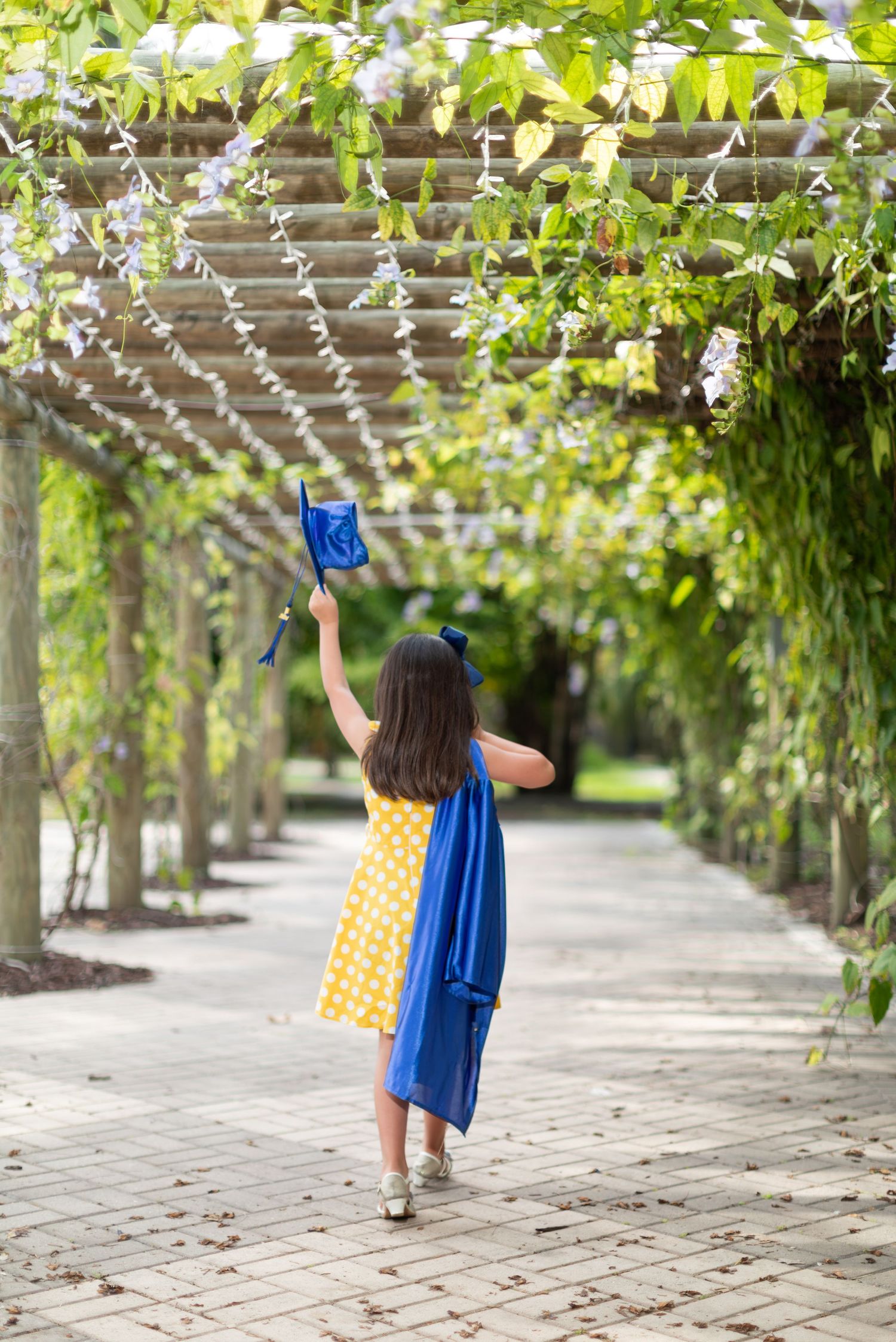 A graduate holds their cap and diploma aloft while walking down a scenic garden pathway lined with trees and vines.