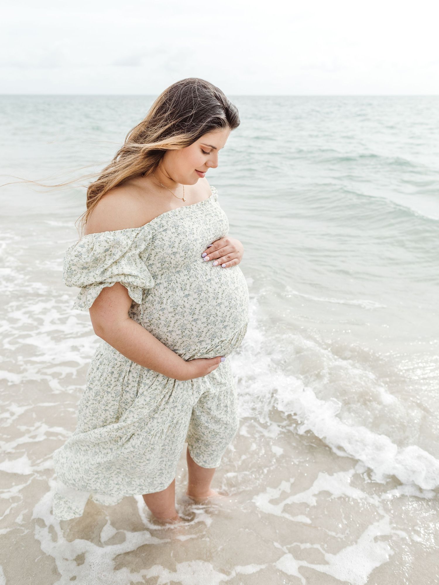 A person wearing a floral dress stands in gentle ocean waves, cradling their pregnant belly on a peaceful beach.