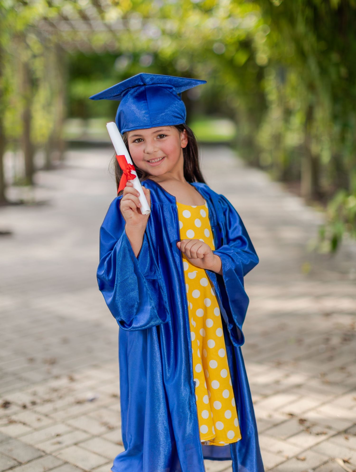 Young graduate in bright blue cap and gown poses outdoors while holding diploma and wearing polka dot dress.