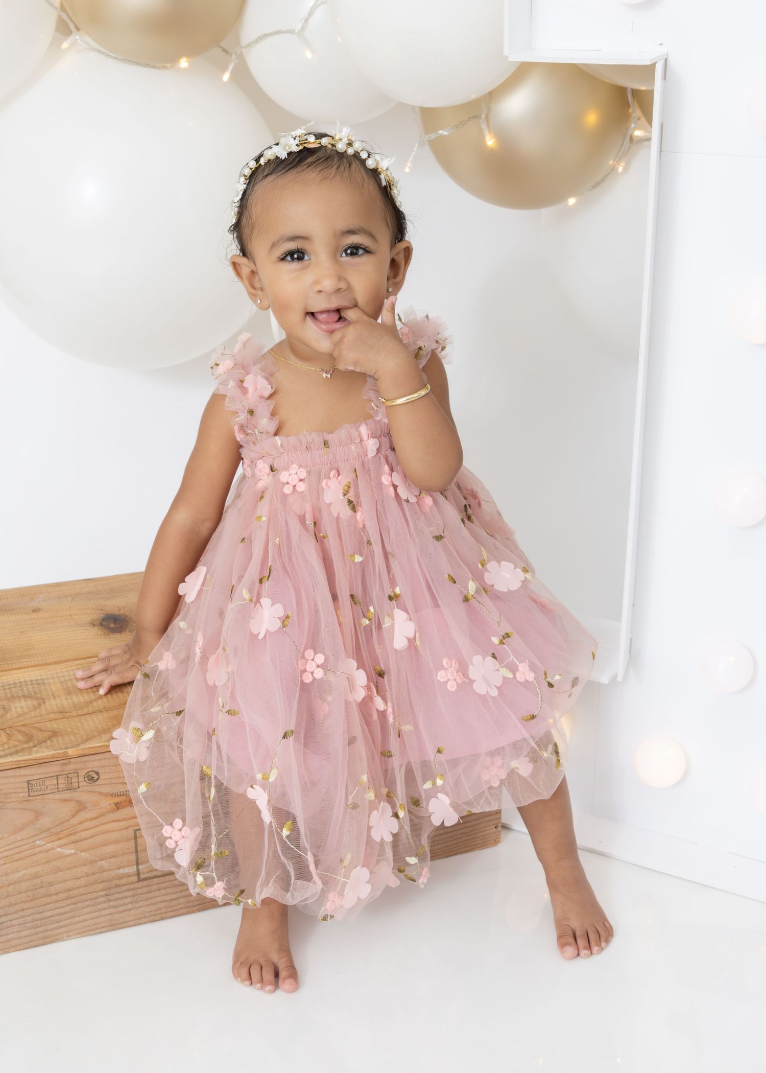 A baby in a pink tulle dress with floral appliques sits on a wooden floor surrounded by gold and white balloons.