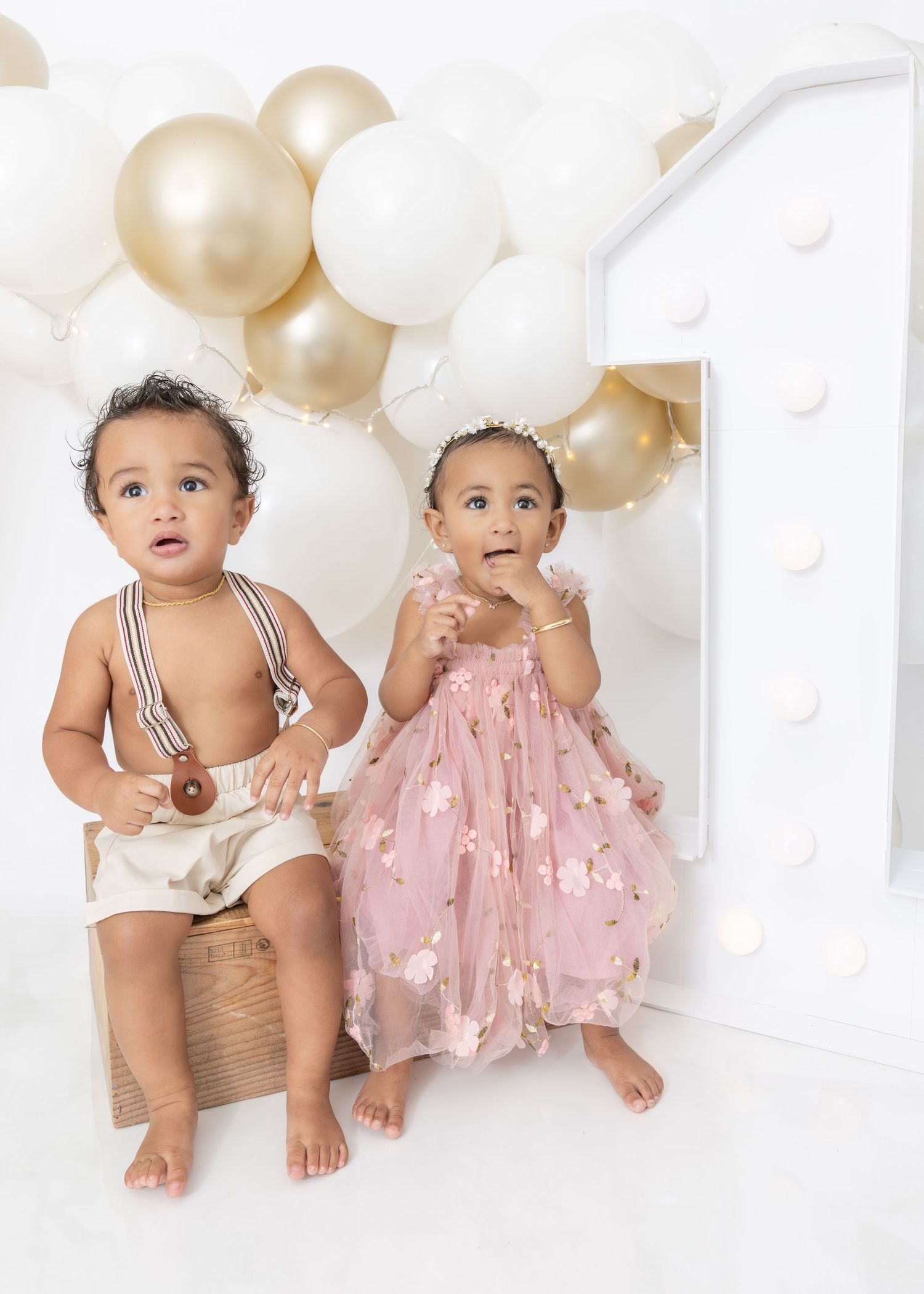 Two babies in matching striped and pink outfits sit together against a white background with gold and white balloons.