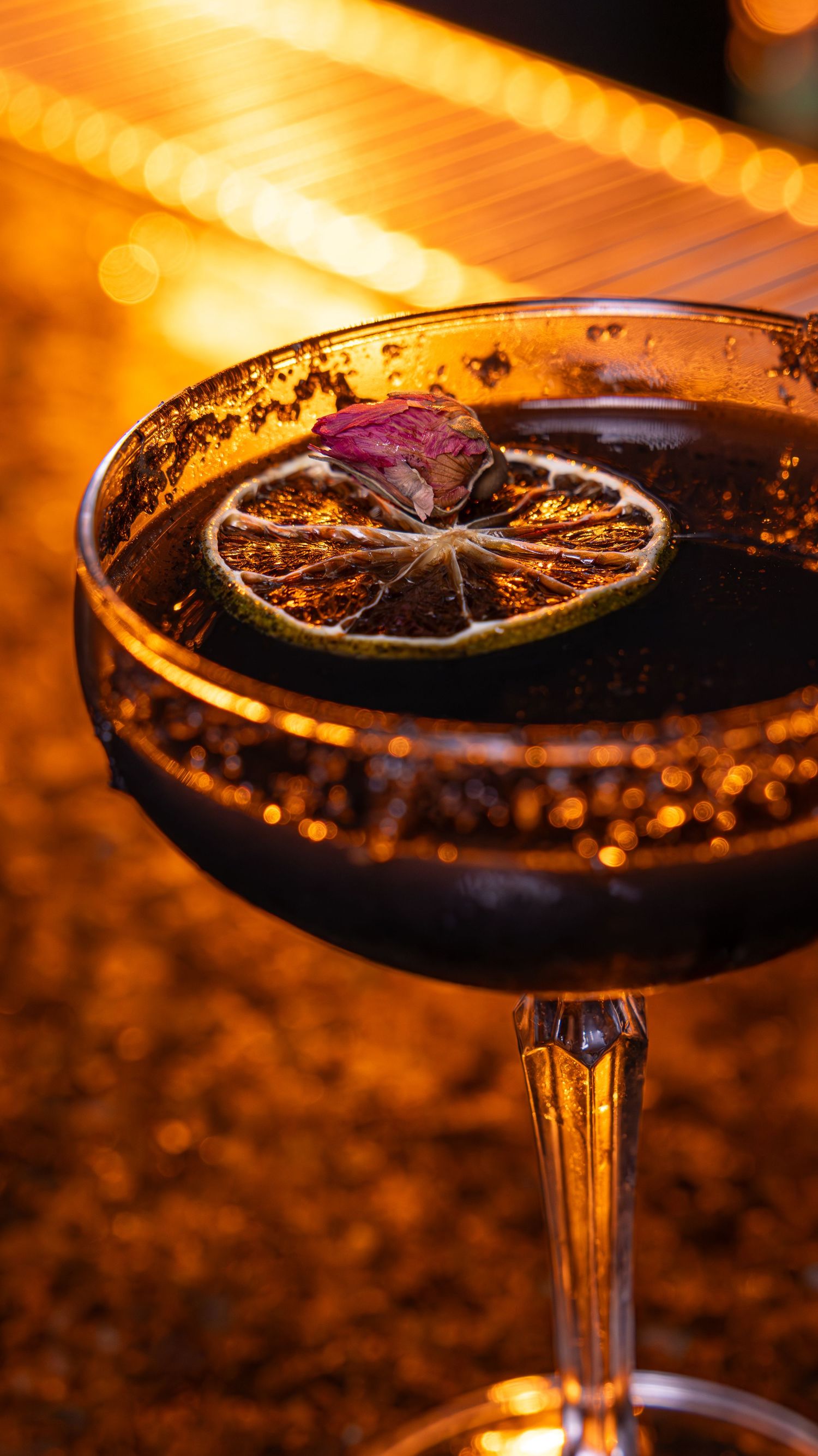 A dark cocktail glass garnished with a dried citrus wheel glowing warmly in amber lighting at a bar.