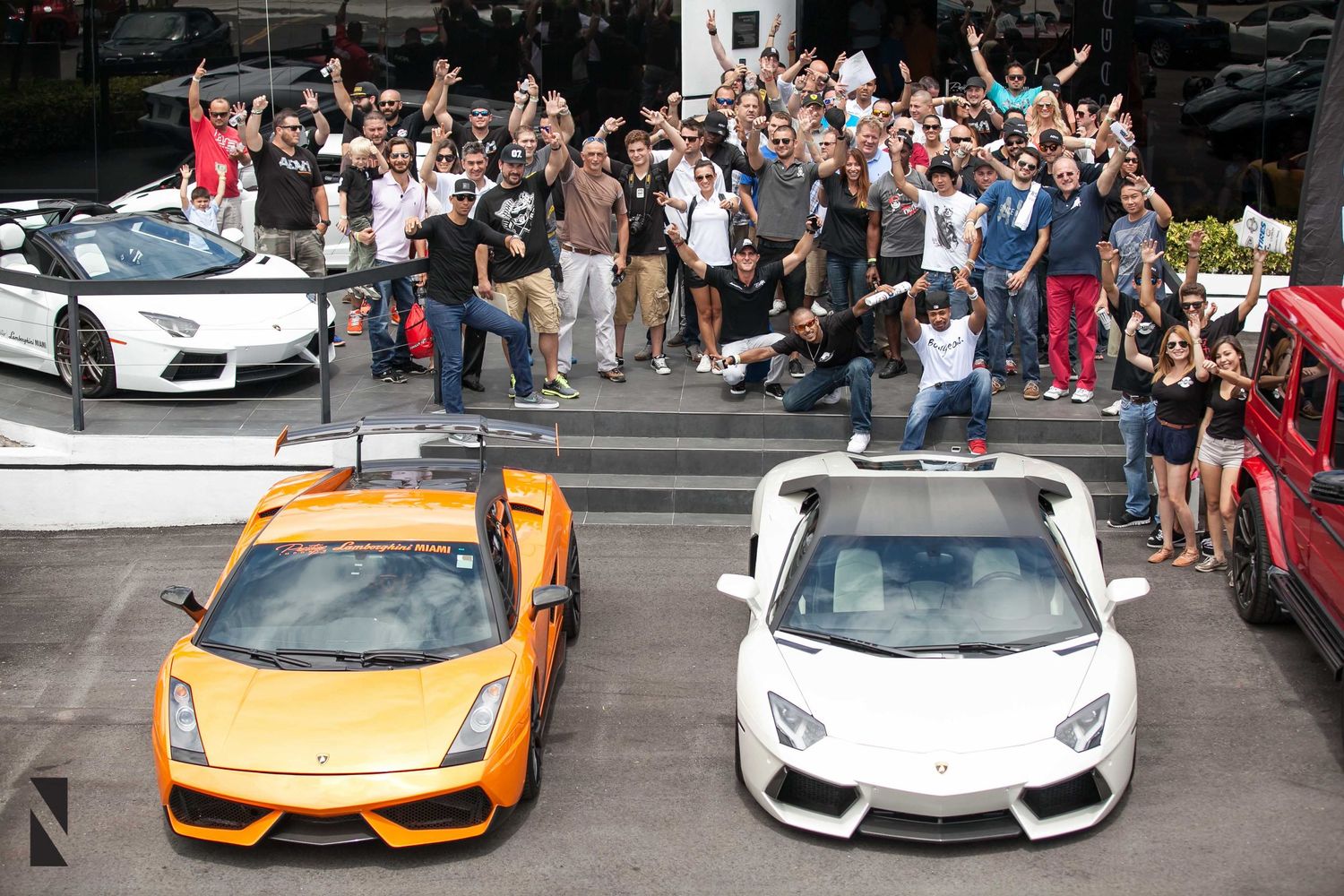 A crowd gathers around two Lamborghini sports cars, one orange and one white, parked on display at an event.