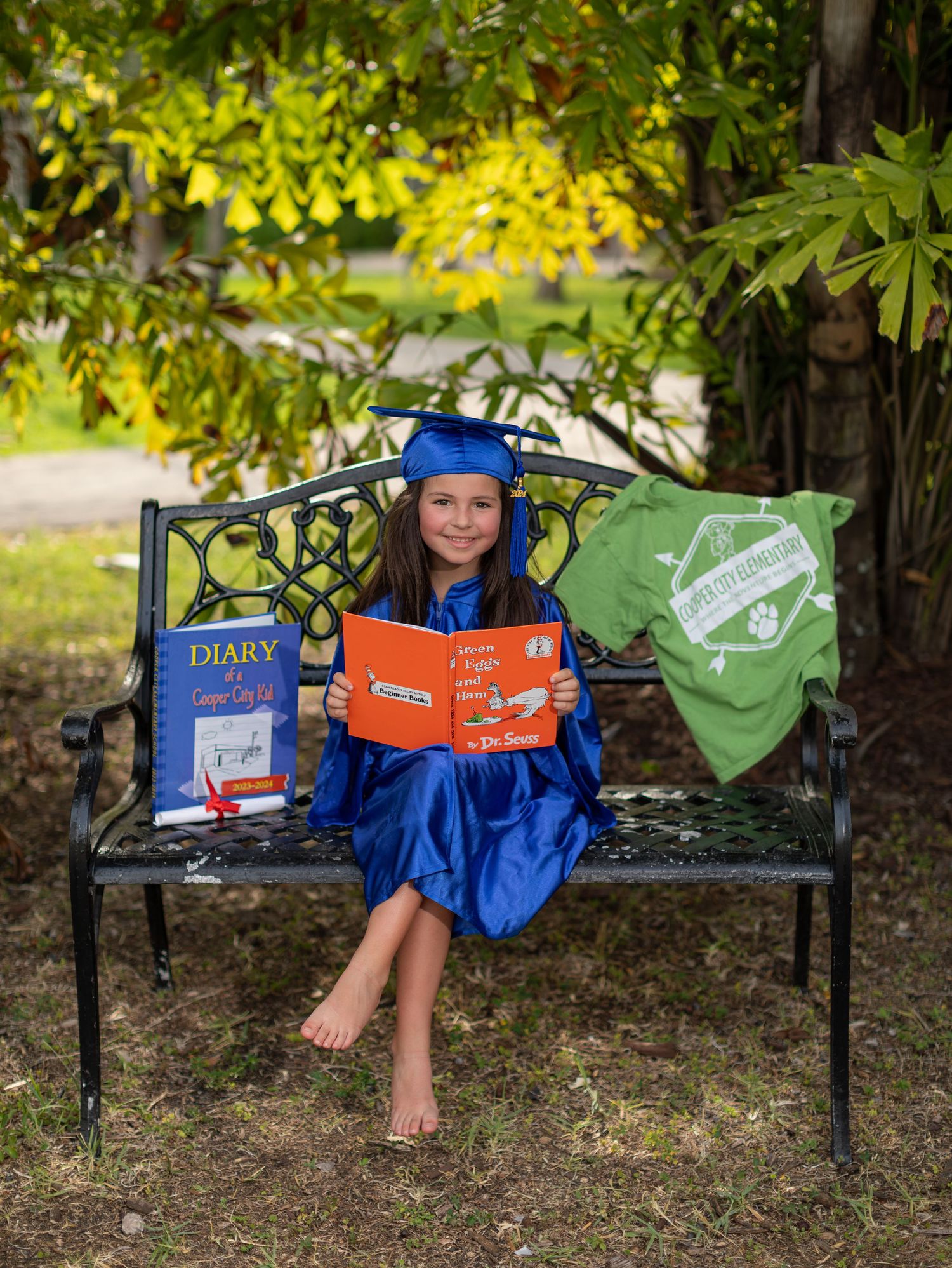 Graduate in blue cap and gown sits on garden bench with books and keepsake items in outdoor setting with autumn foliage.