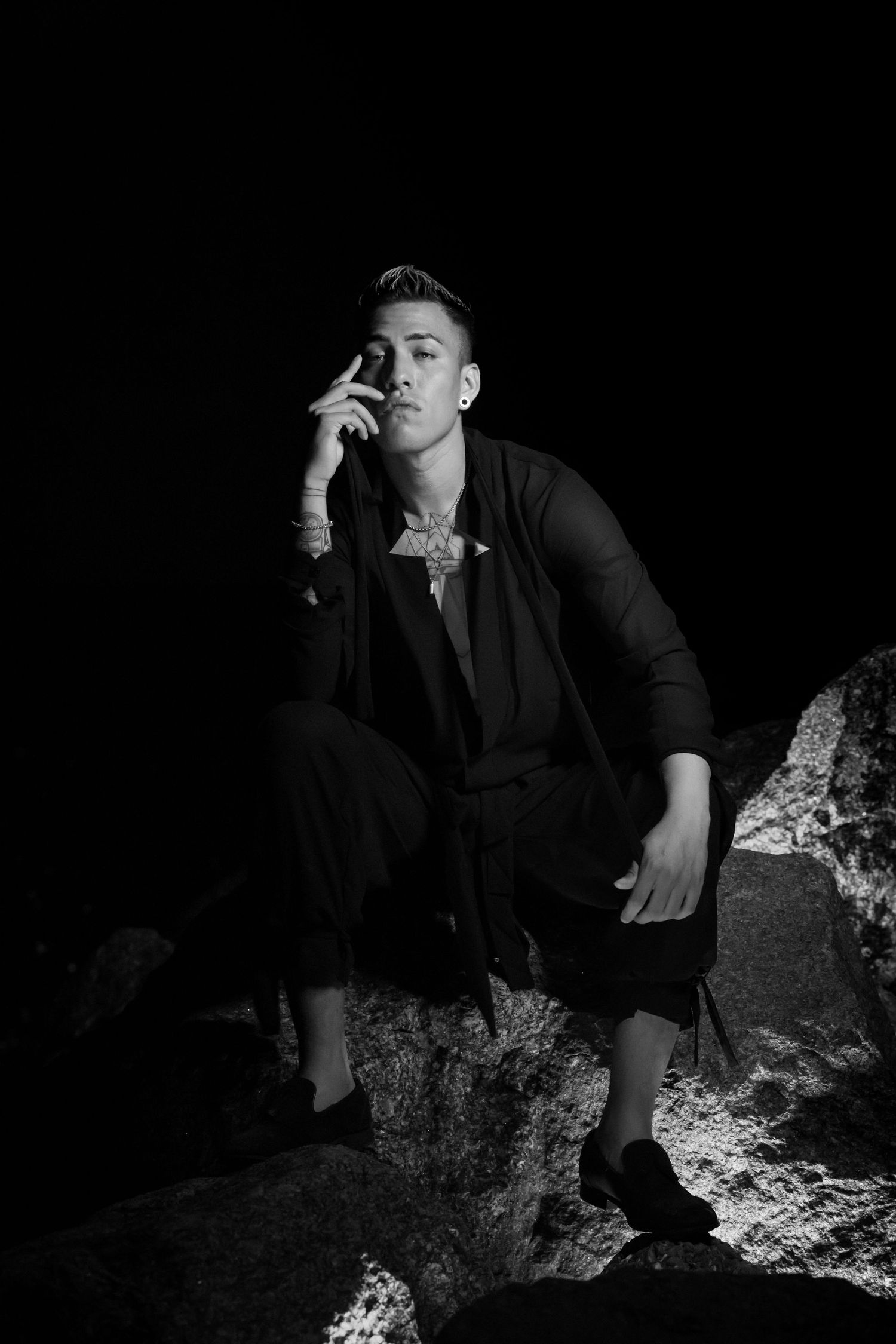 Dramatic black and white photo of figure seated on rock wearing dark formal attire.