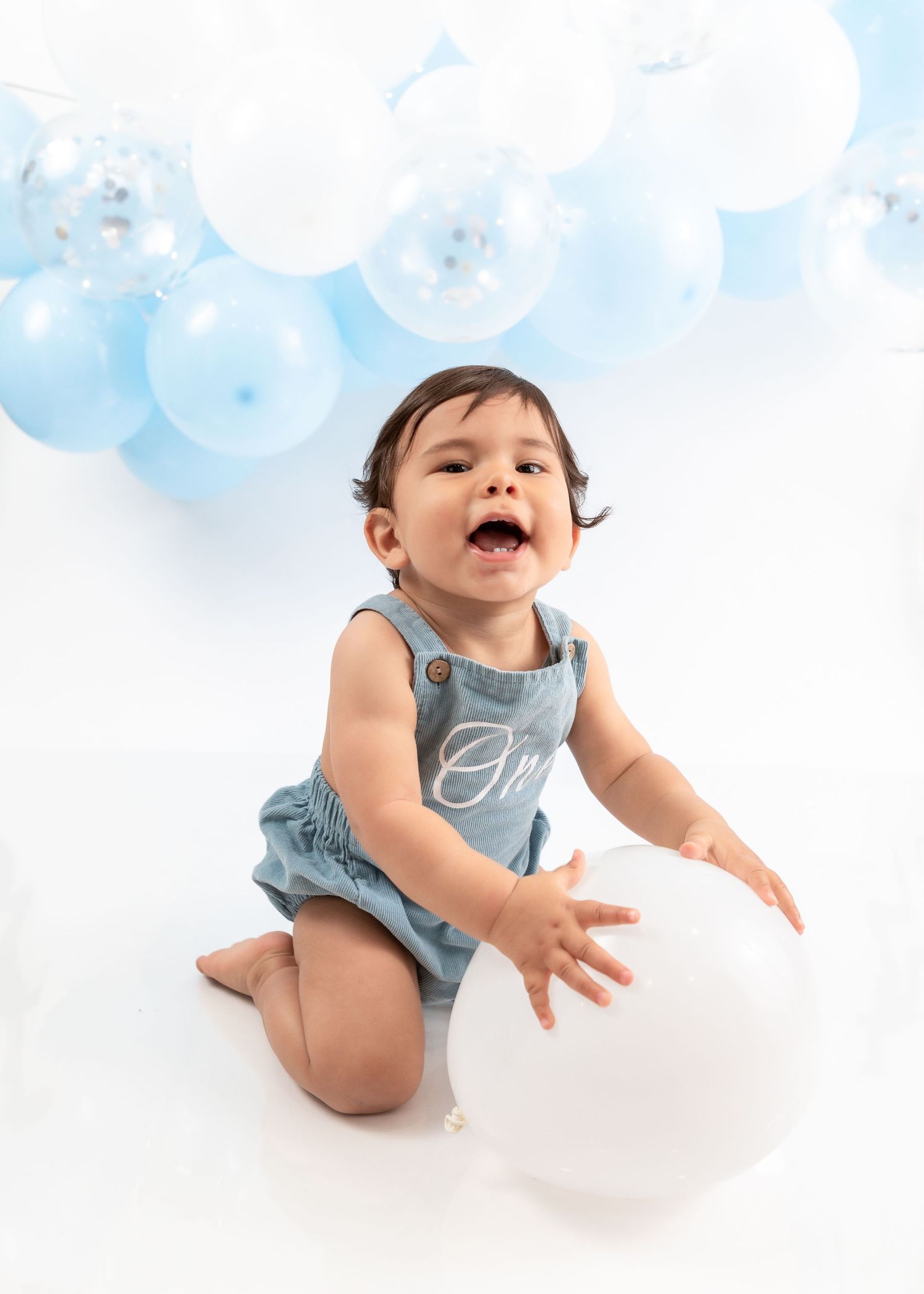 Baby playing with balloons in light blue outfit during first birthday photoshoot.