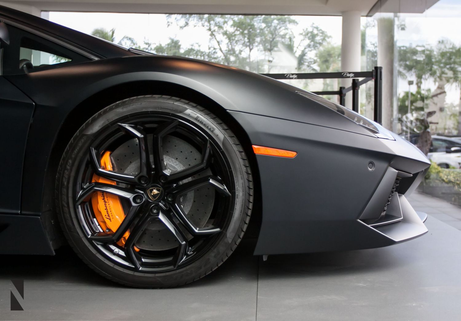 Close-up view of a gray Lamborghini Aventador's front wheel showing orange brake caliper and black rim details.