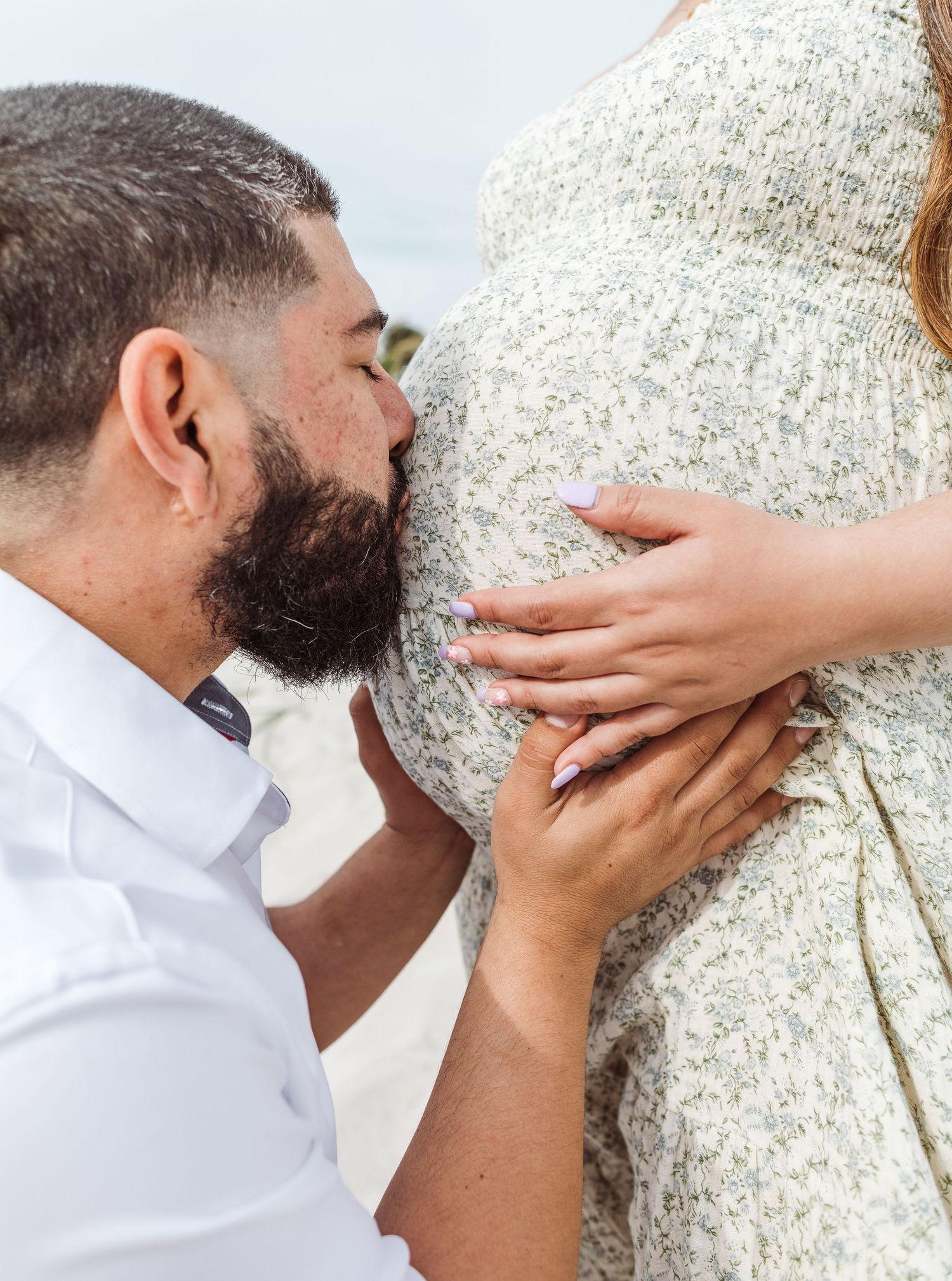 A person in a white shirt kisses and holds a pregnant belly while wearing a full beard.