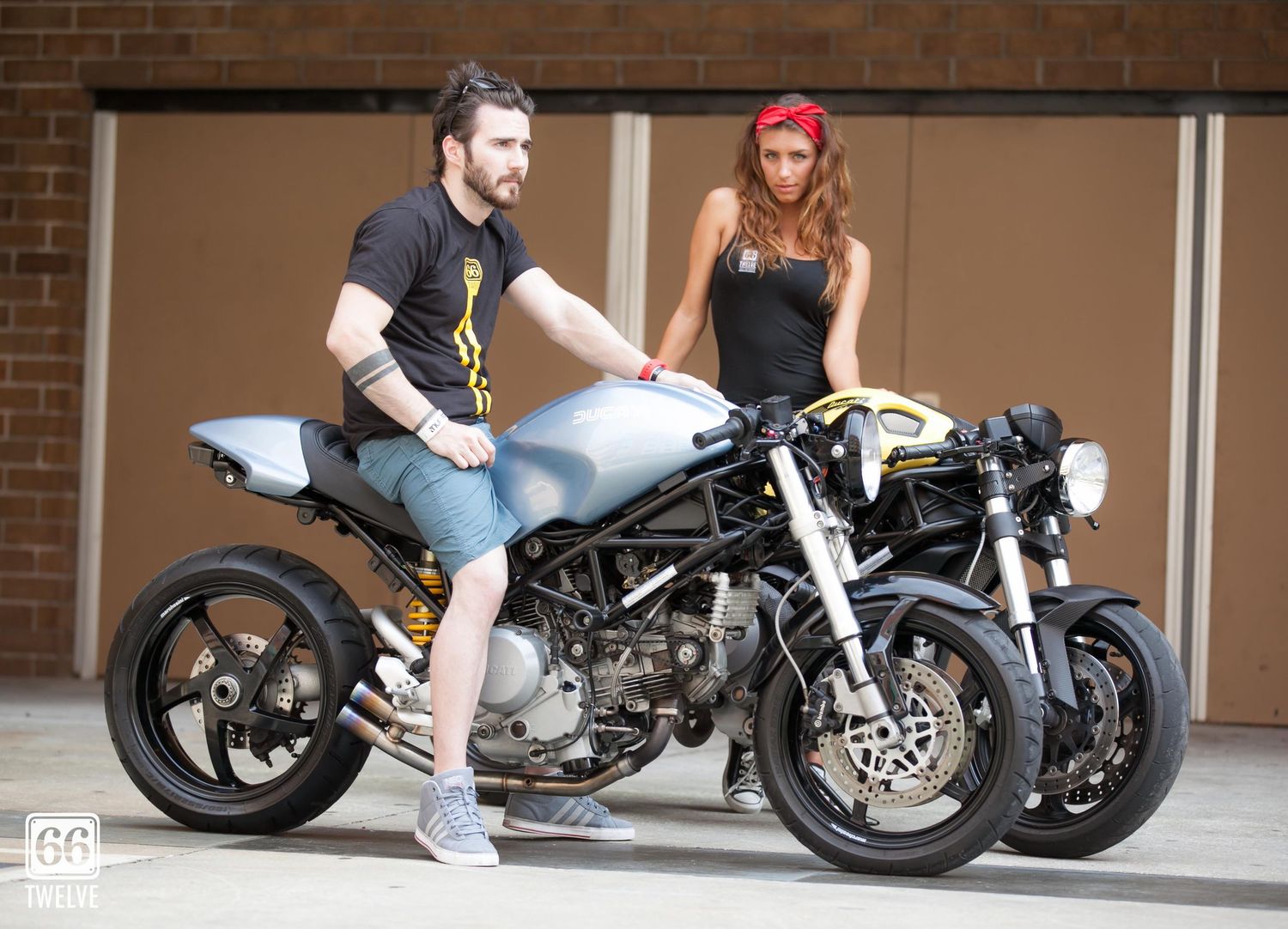 Two people pose with a custom silver cafe racer motorcycle against a brick wall backdrop.