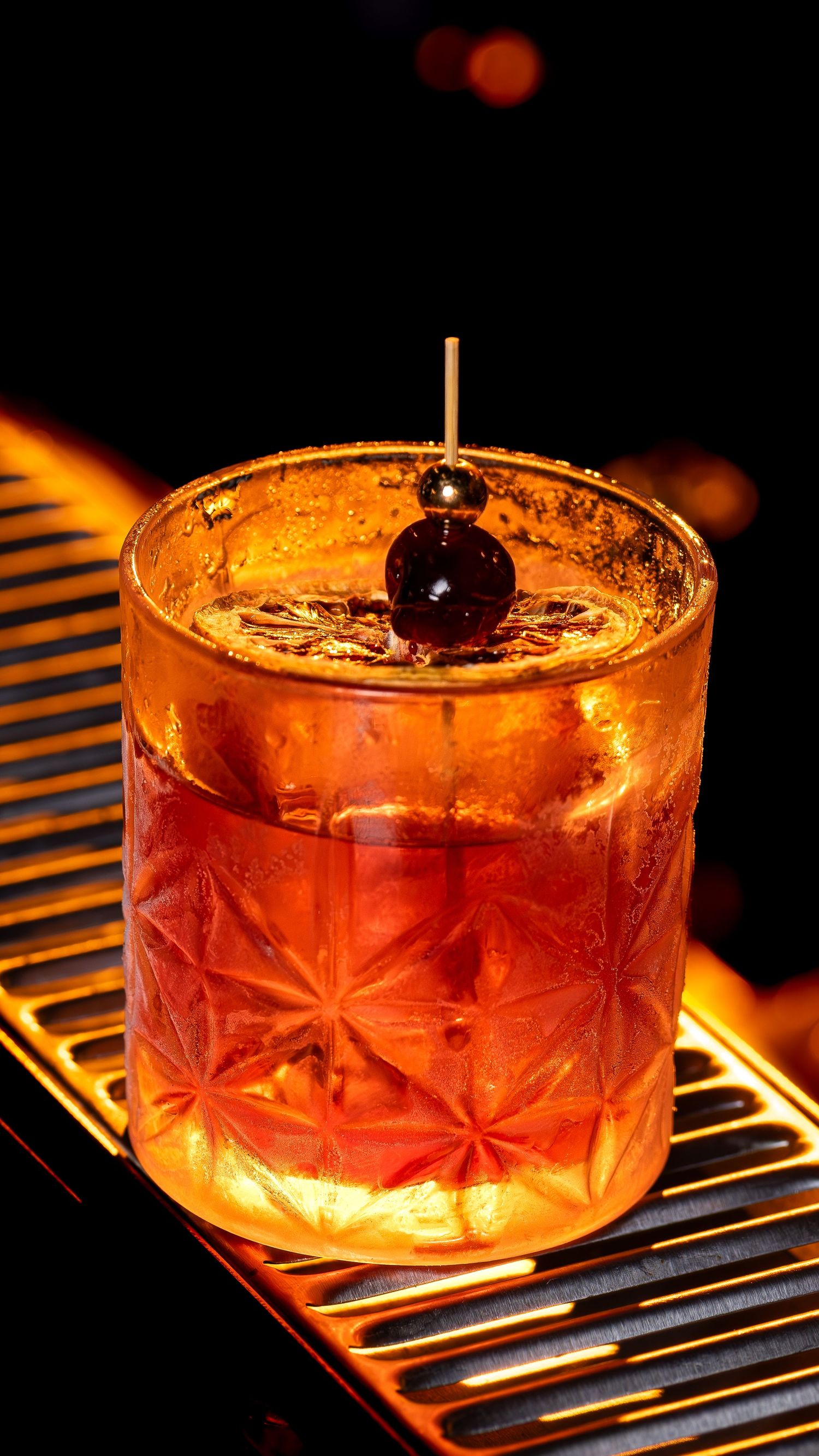A classic cocktail served in a crystal cut glass with a dark cherry garnish sits glowing on a metal bar rail.