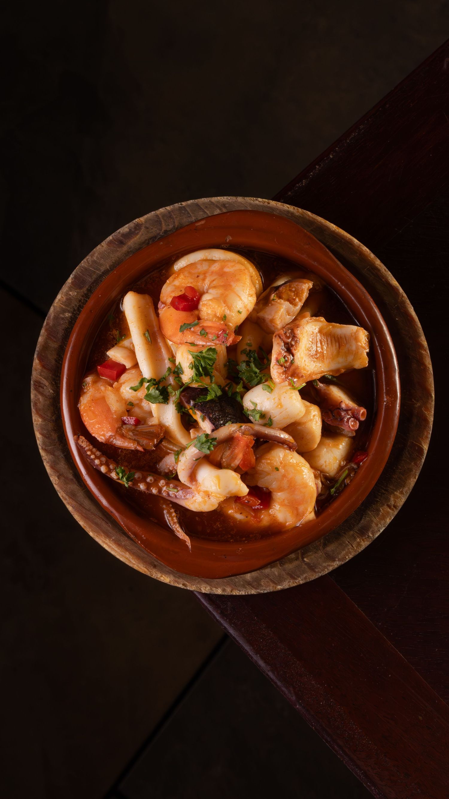 Overhead view of shrimp and seafood in a rustic terra cotta bowl with herbs and broth.