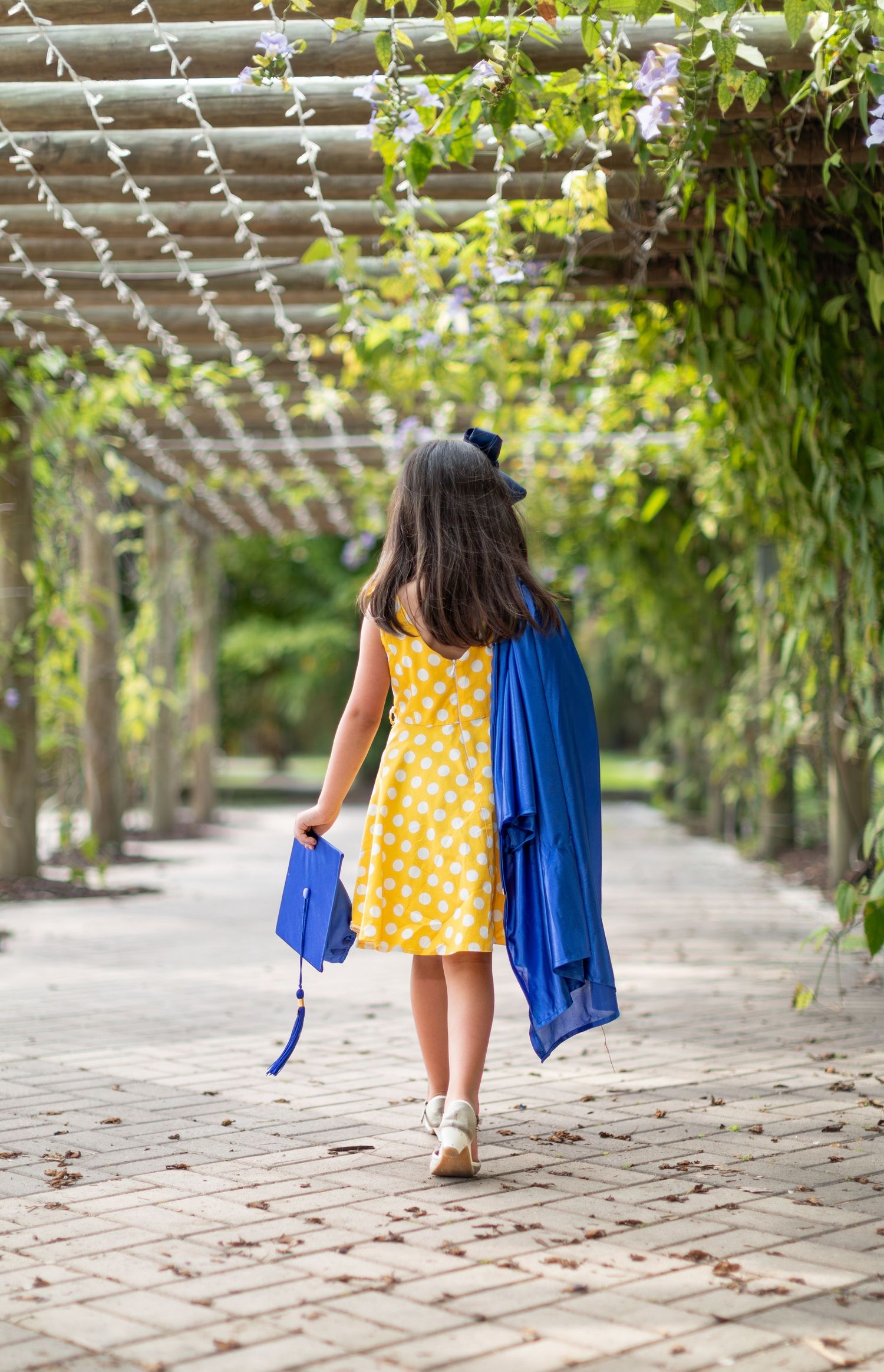 Figure in yellow polka dot dress walks under garden pergola carrying blue coat on stone pathway.