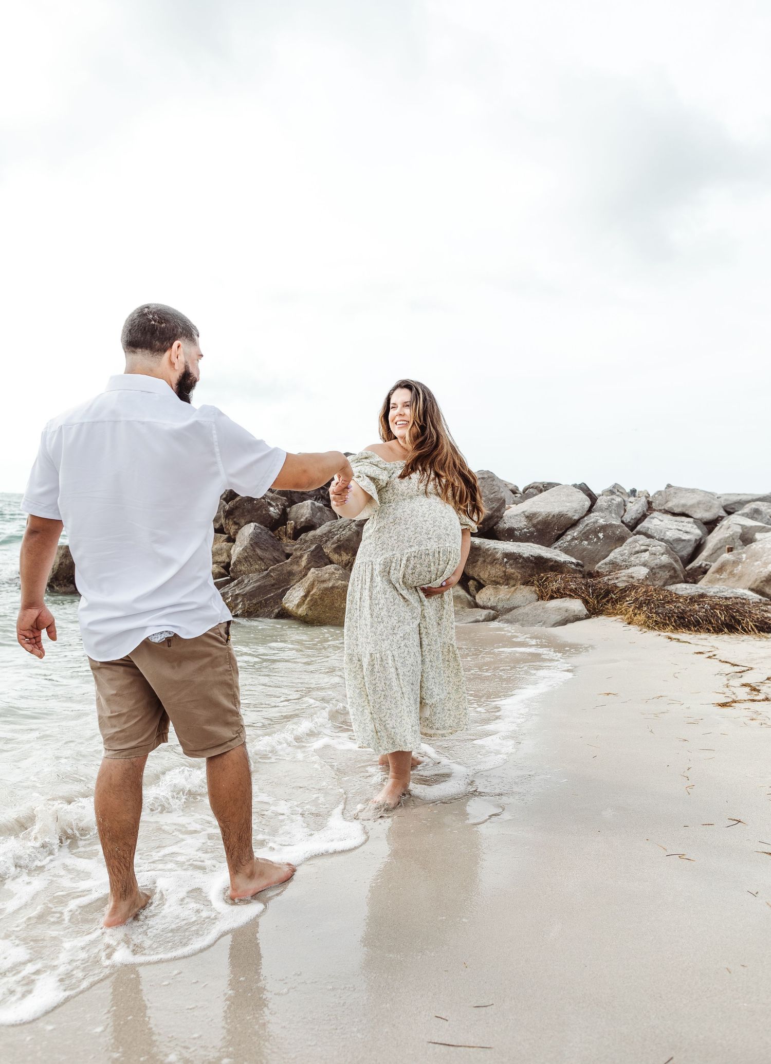 Pregnant couple in casual attire dance together on a rocky beach shoreline during an overcast day.
