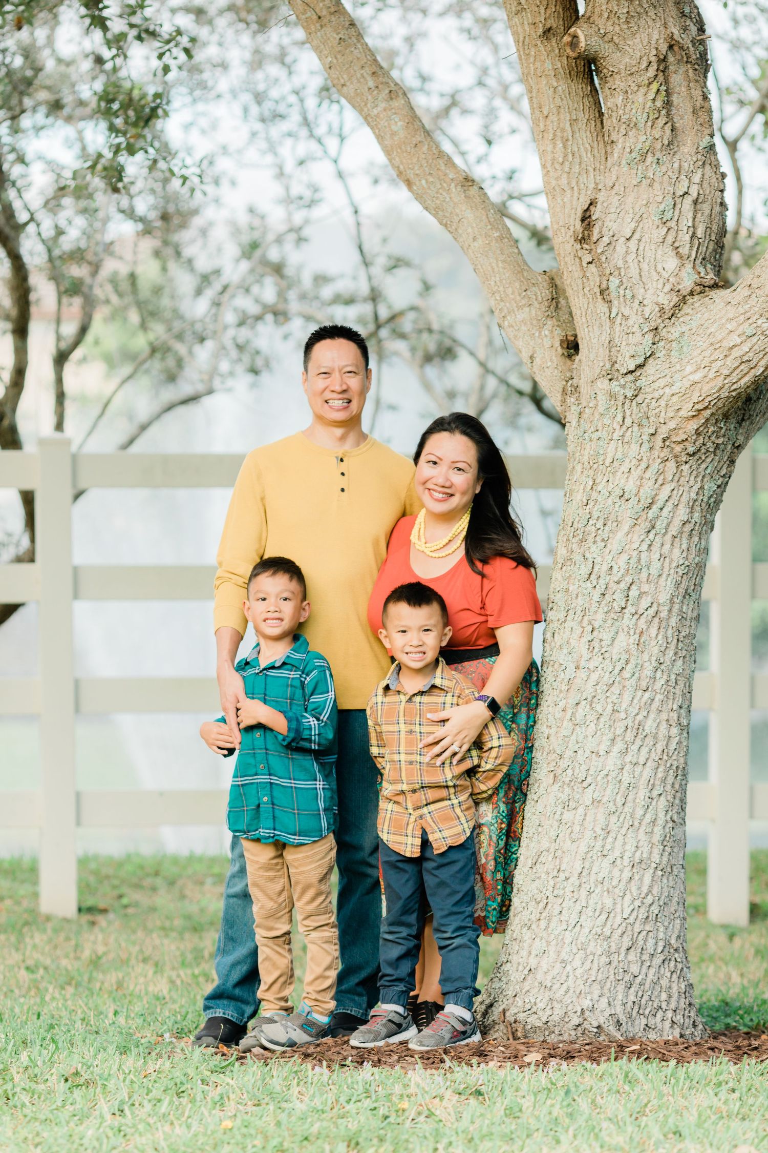 Family portrait of four people standing together by a tree with white fence in background.