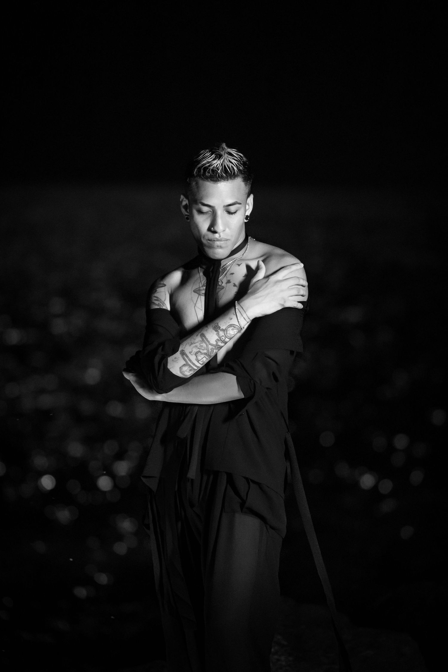Artistic black and white portrait of person in dark attire posing with crossed arms against blurred lights background.