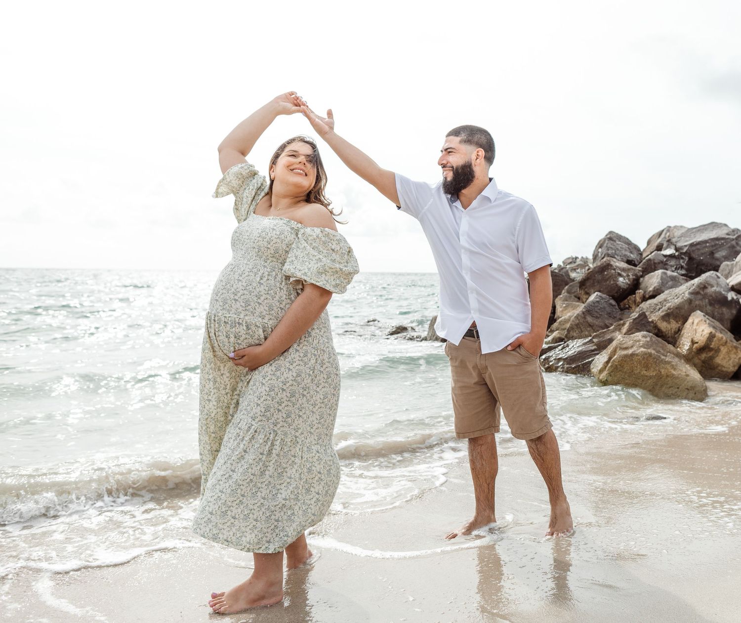 Two people dance and smile together on a rocky beach with ocean waves in the background.
