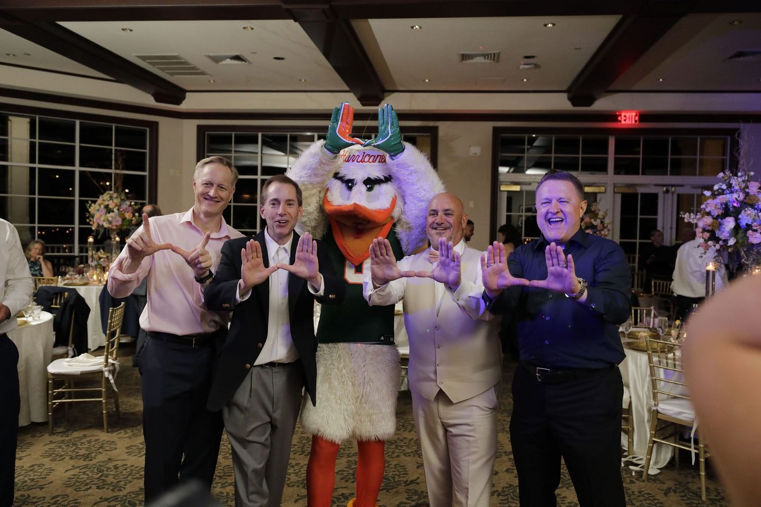 A group of people pose with a costumed mascot character at an indoor event with decorative lighting and floral arrangements.