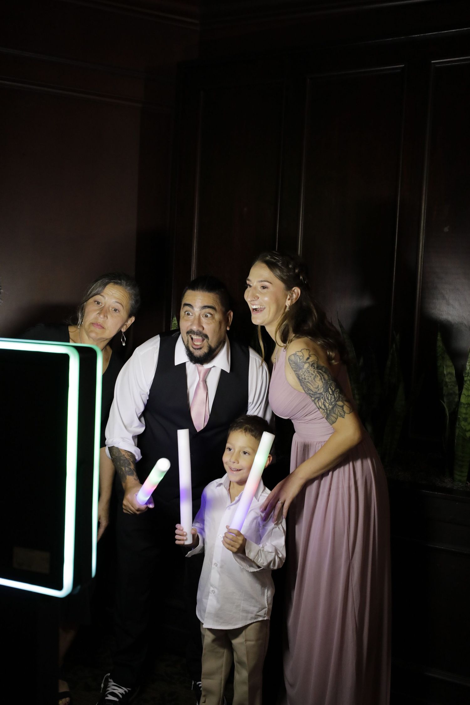 A family of three poses with glowing light sticks at a dark reception venue, with a neon-lit photo booth in the background.