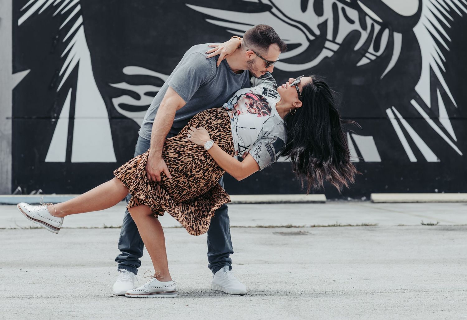 A playful couple performs a dramatic dip dance move against an urban art wall.
