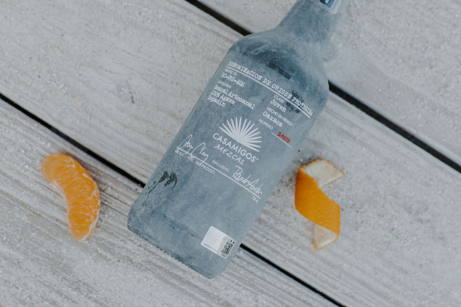 A bottle of Casamigos tequila lies on a gray wooden surface with orange citrus wedges arranged beside it.